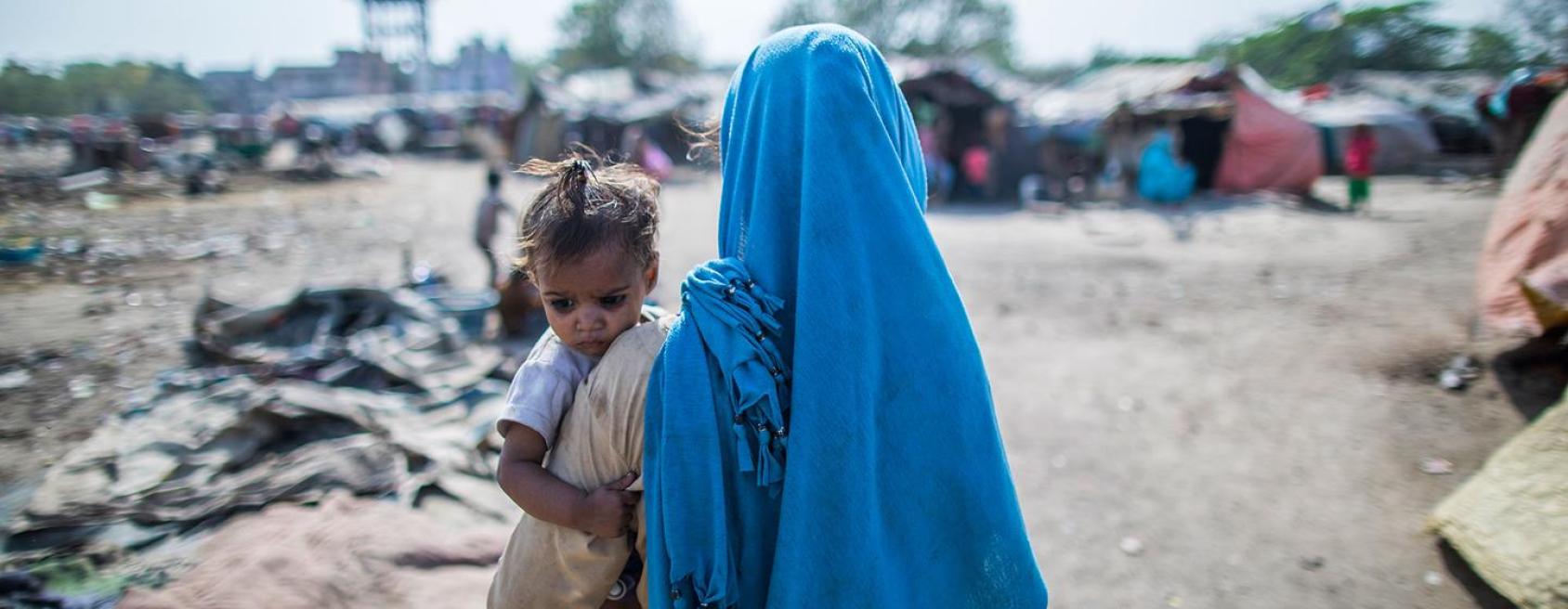 Une femme portant un long voile bleu et tenant un jeune enfant dans les bras est photographiée de dos, debout au milieu d’un camp de réfugiés.