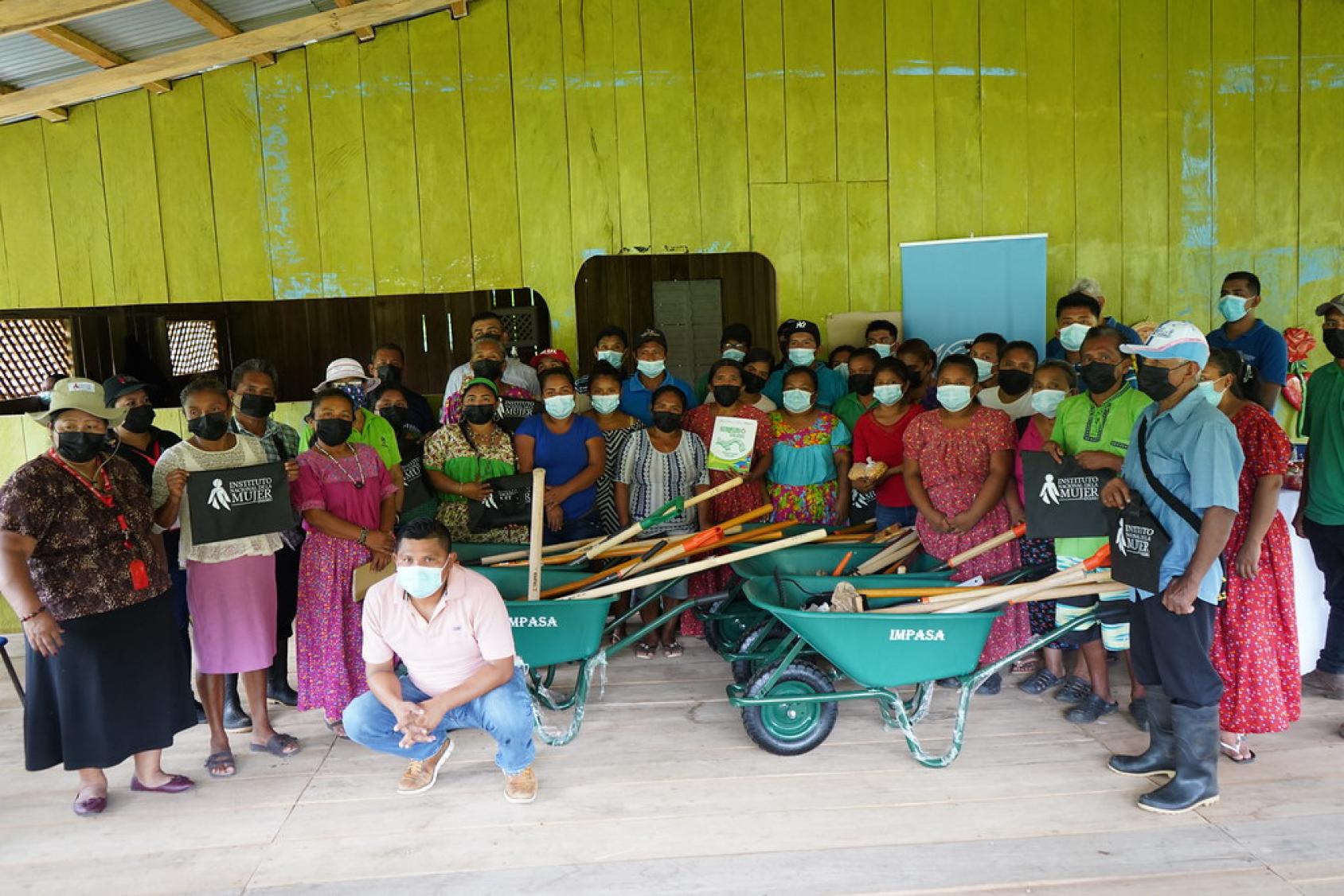 Group picture of Naso Tjër Di women and men wearing face masks and some of them are holding wheelbarrows and other farming tools and implements.