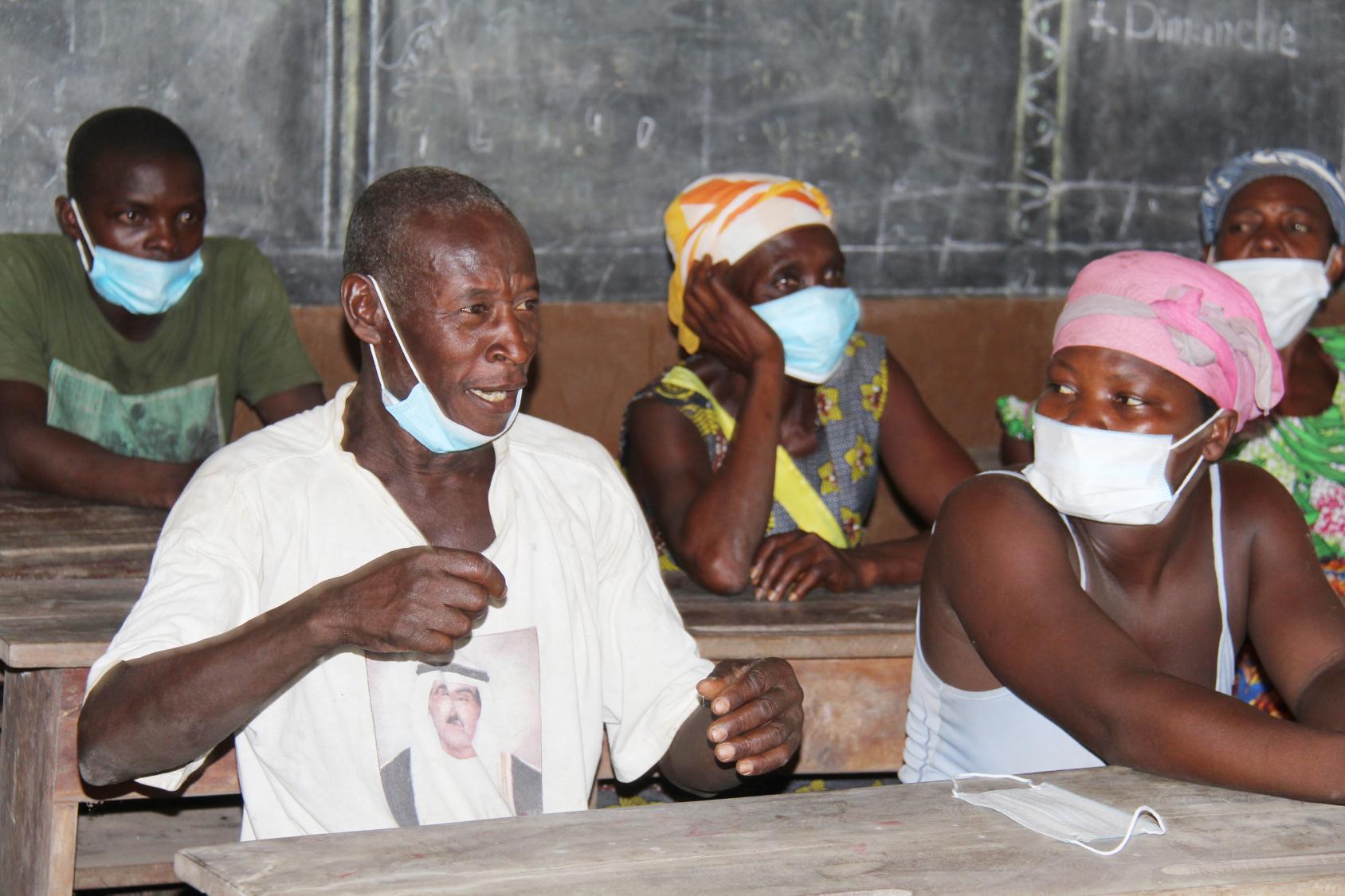 An older man is sitting at a wooden table in a classroom and speaking with a face mask pulled down under his chin. Several people, all wearing face masks, are sitting next to him and behind him.
