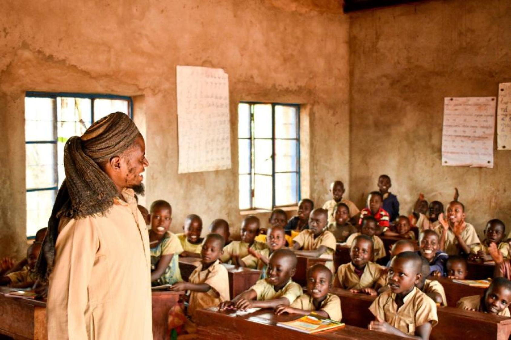Un homme portant un turban et un habit traditionnel burundais de couleur beige fait classe à de jeunes garçons dans une école.