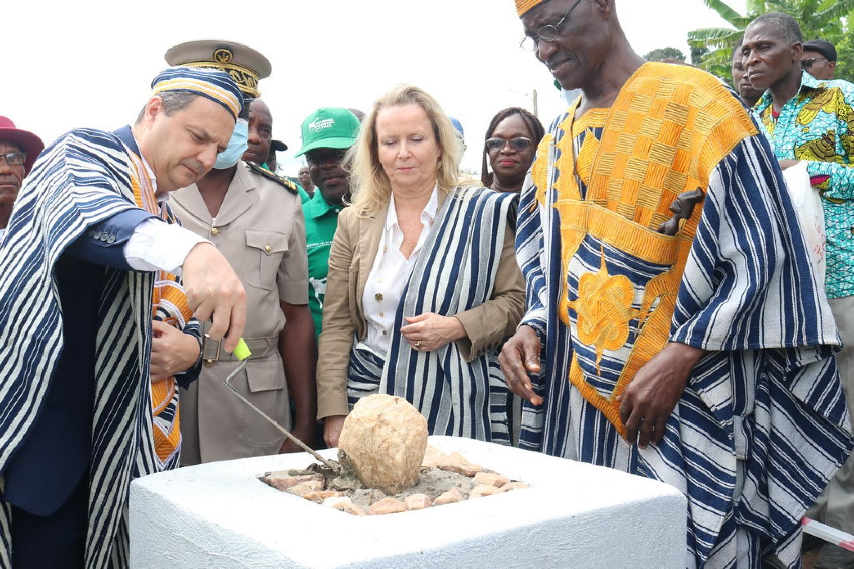 Le Coordonnateur résident des Nations Unies Philippe Poinsot pose la première pierre d'un moulin multifonctionnel. Vêtu d'un habit traditionnel ivoirien, il se tient à côté de la Représentante de l'UNESCO en Côte d'Ivoire et du Chef du canton de Biankouma, lui aussi vêtu d'un habit traditionnel.