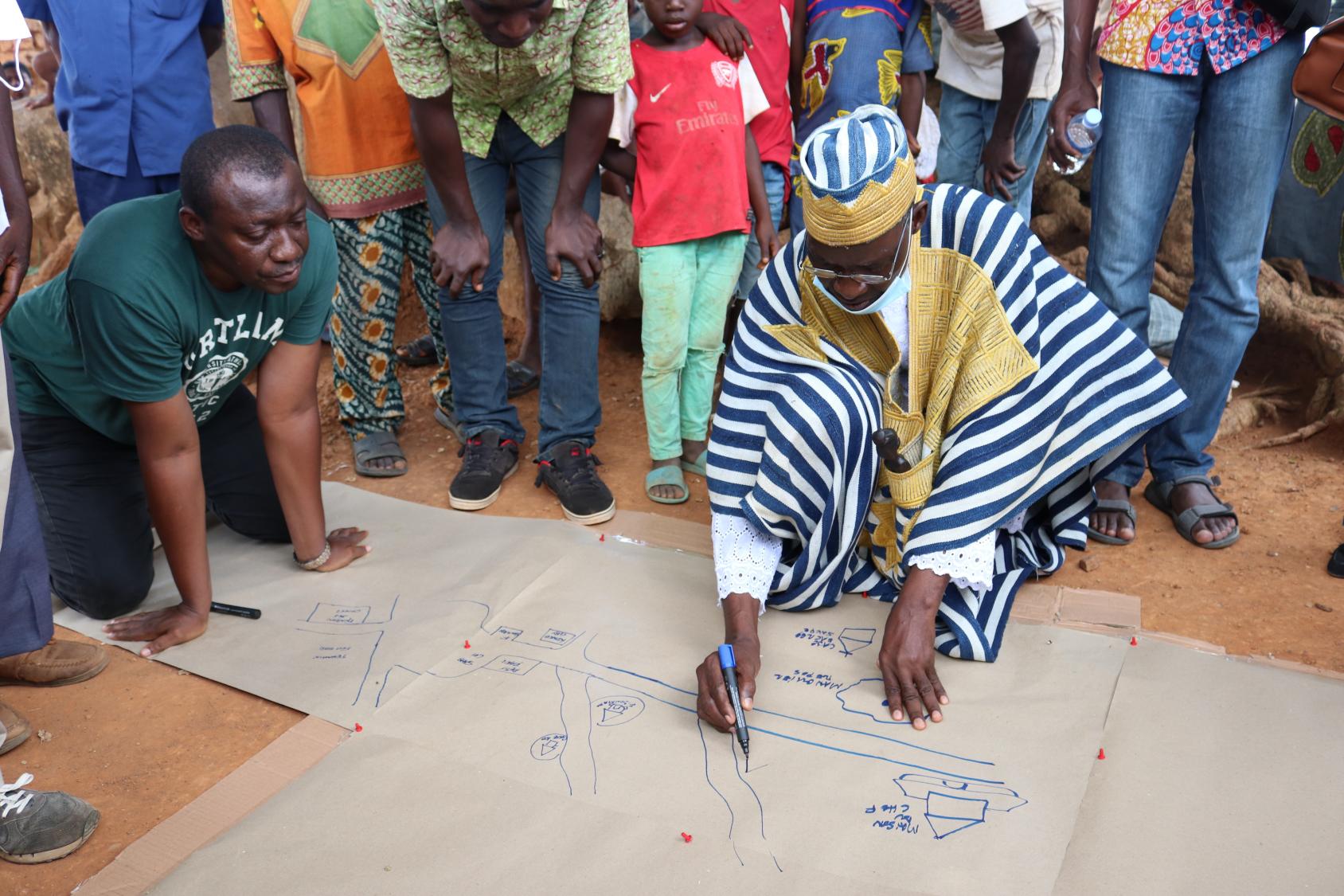 Un homme vêtu d'un habit traditionnel ivoirien est accroupi au sol, entouré de plusieurs personnes, et réalise un inventaire sur une large feuille de papier.