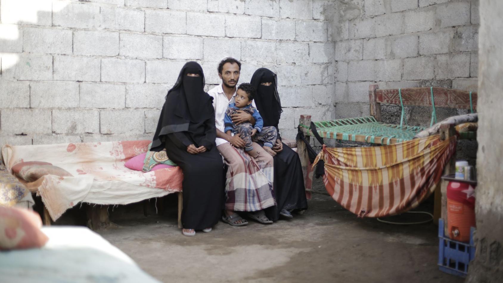 一个男人和两个戴着黑纱的女人坐在一起，孩子坐在他的腿上，在一堵砖墙前。