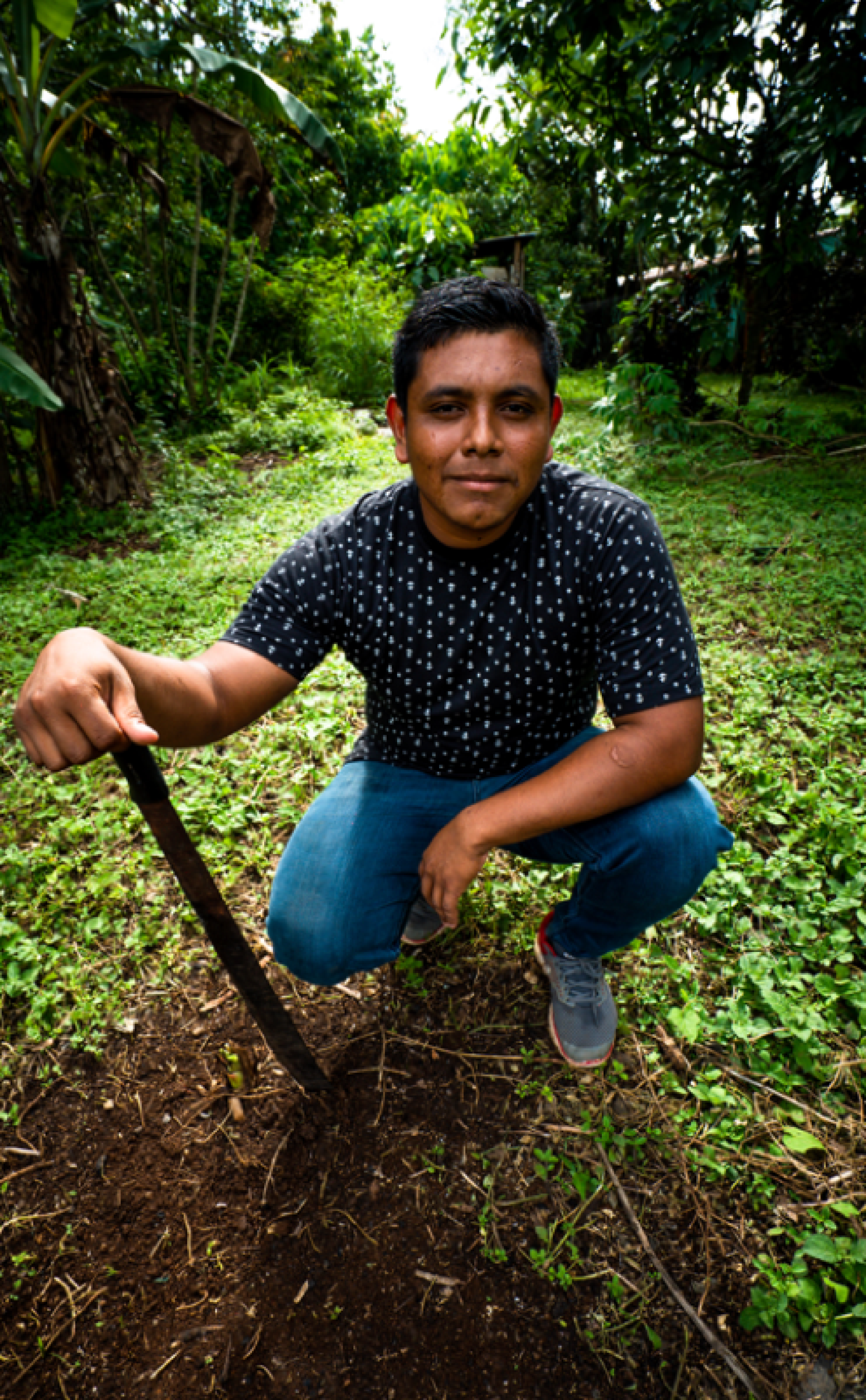 A young man bends down near a shovel in a green patch field with trees.
