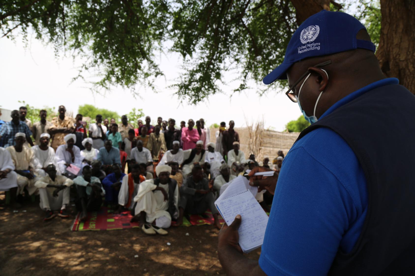 A Al Fado, une localité d'Assalaya, dans le Darfour oriental, un membre du personnel de maintien de la paix des Nations Unies s’adresse à un groupe de personnes assises à même le sol, à l’ombre d’un arbre, tout en lisant des notes manuscrites dans un petit carnet.