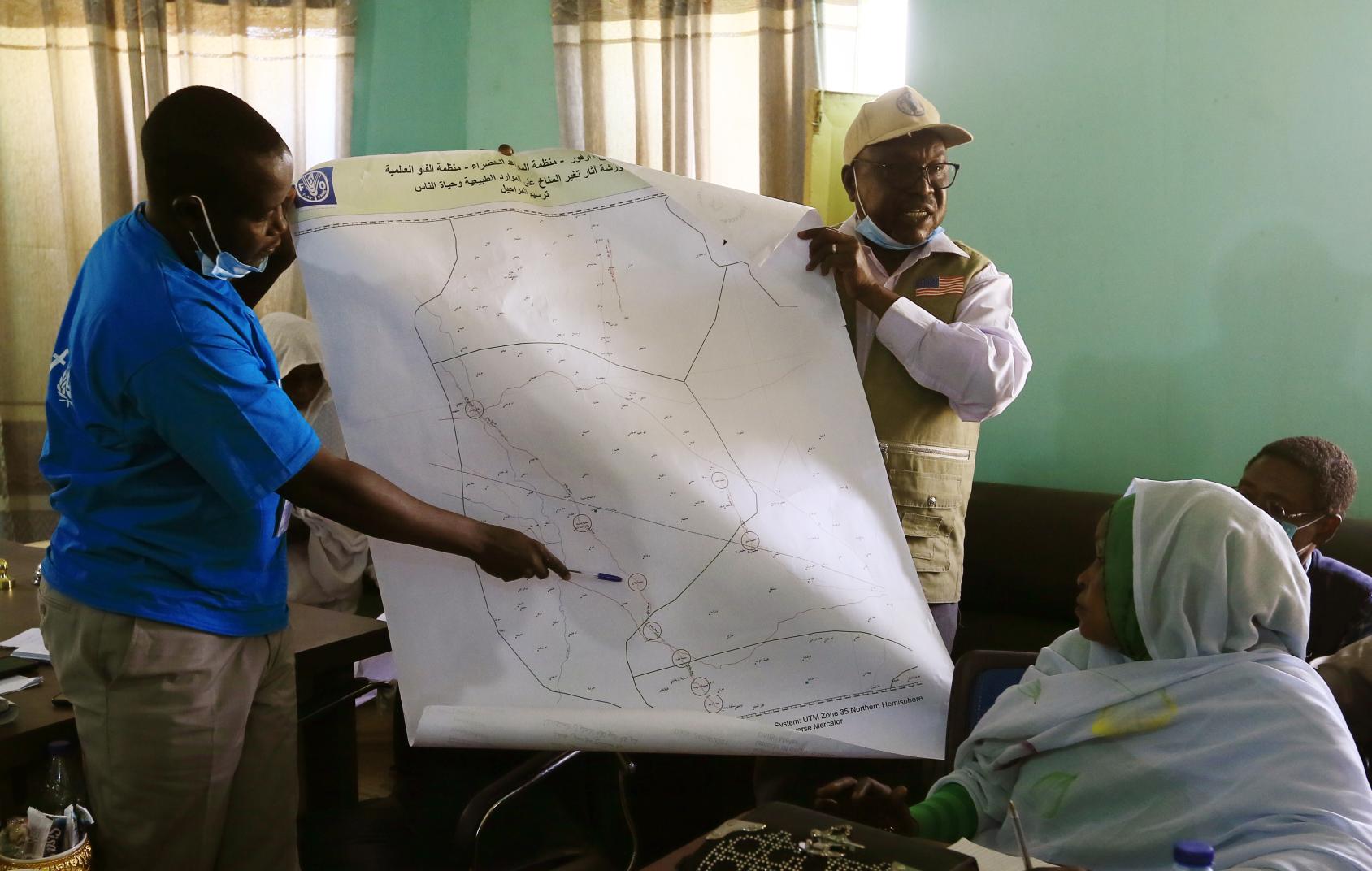 Deux hommes expliquent à des personnes réunies dans une salle où se trouvent les routes migratoires et les foyers de conflit cartographiés par la FAO dans le village de Yassin, dans le Darfour oriental. 