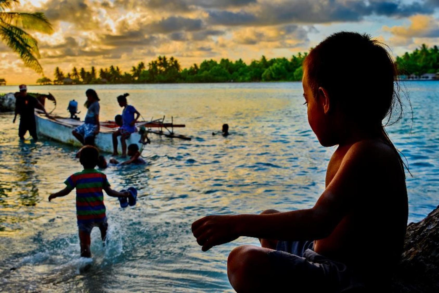Siluetas de niños y niñas jugando en la orilla de la playa en el atardecer, y un niño mira hacia un barco de pesca.