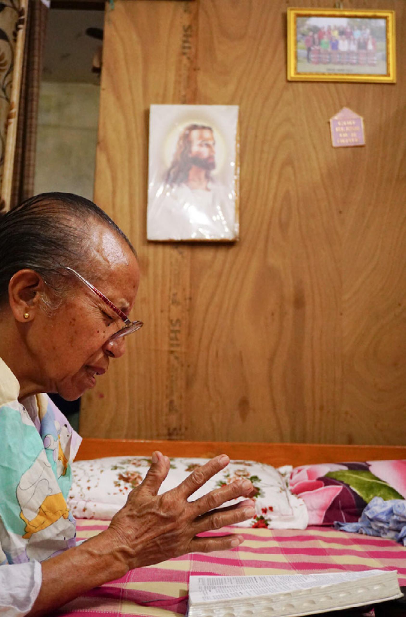 Пожилая женщина молится с открытой Библией на столе.