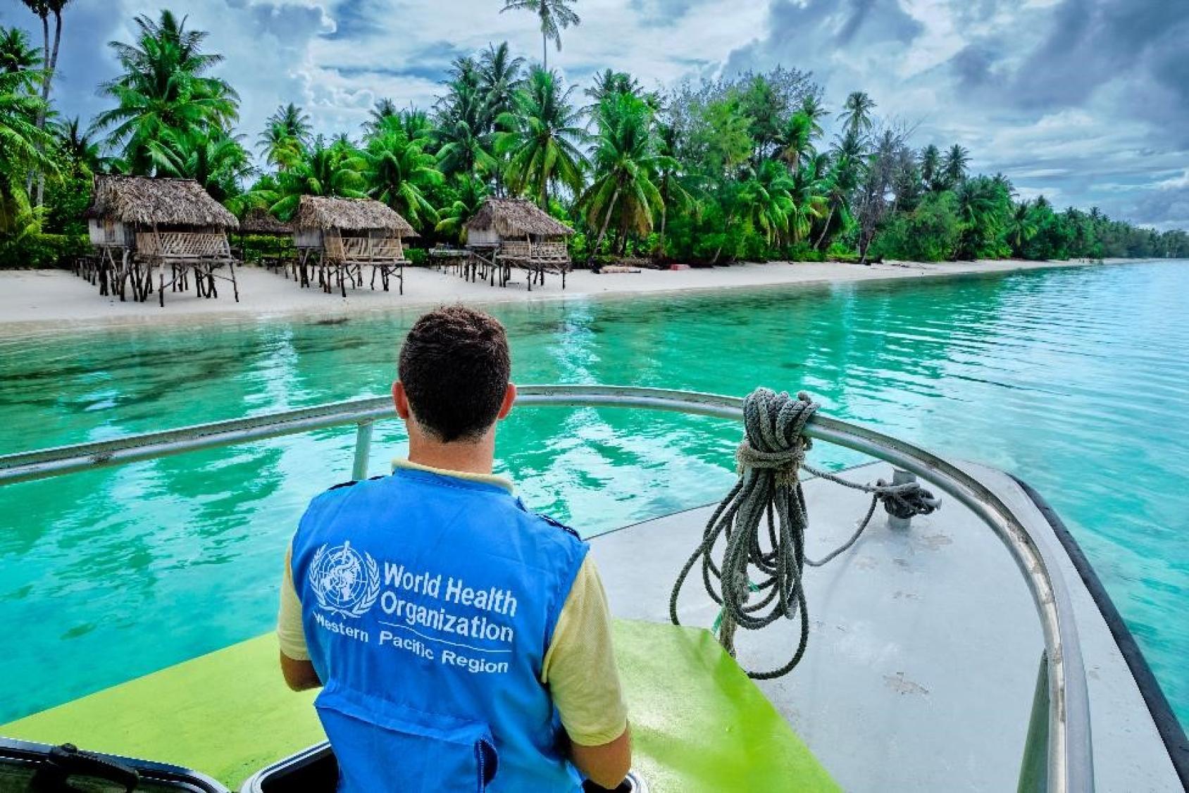 A Kiribati, un membre du personnel de l’OMS portant un gilet bleu navigue à bord d’un bateau dans l’eau turquoise de l’océan, à proximité d’un rivage bordé de palmiers où ont été construites deux rangées de cabanes.