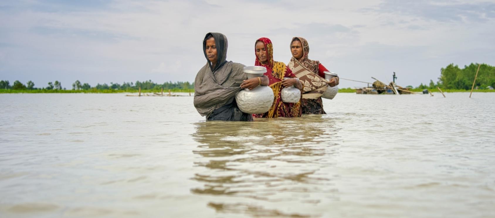 Tres mujeres caminan por un terreno inundado llevando jarras.
