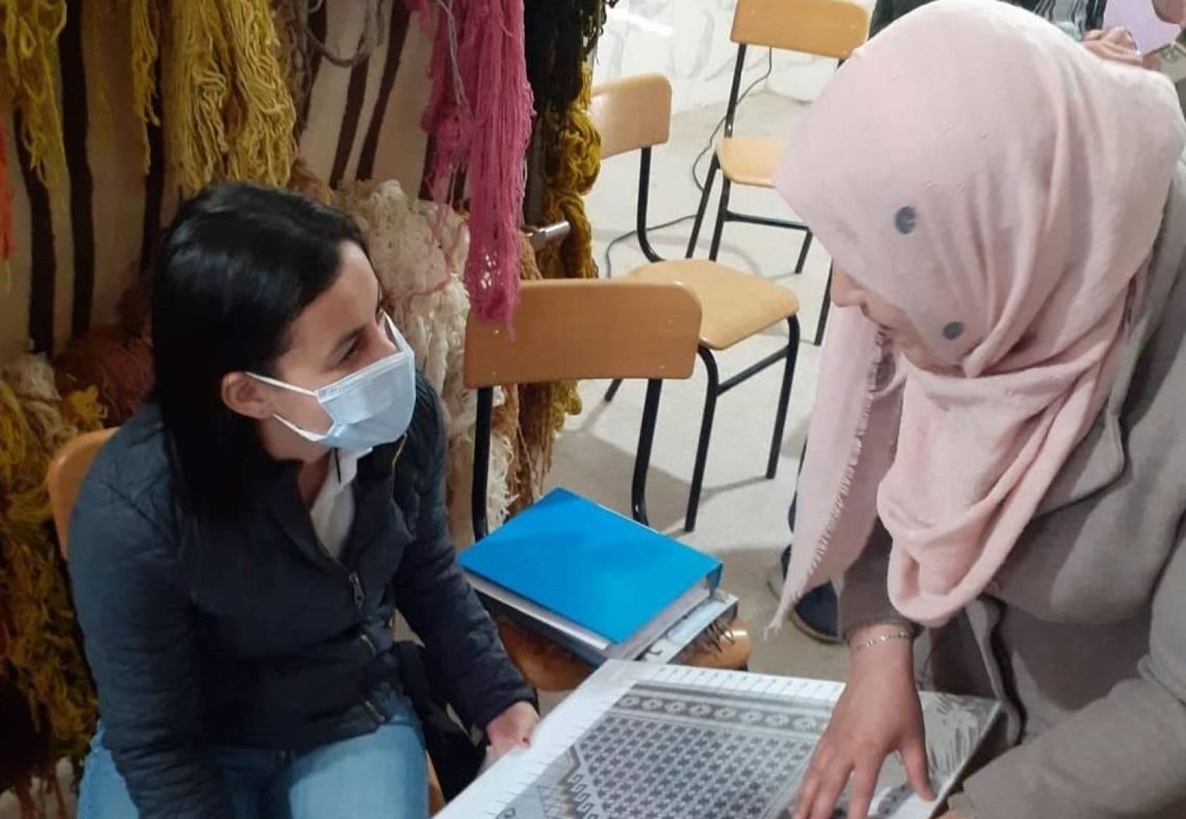 A woman wearing jeans and a face mask talks with a woman wearing a headscarf about a mock-up they both hold in their hands.