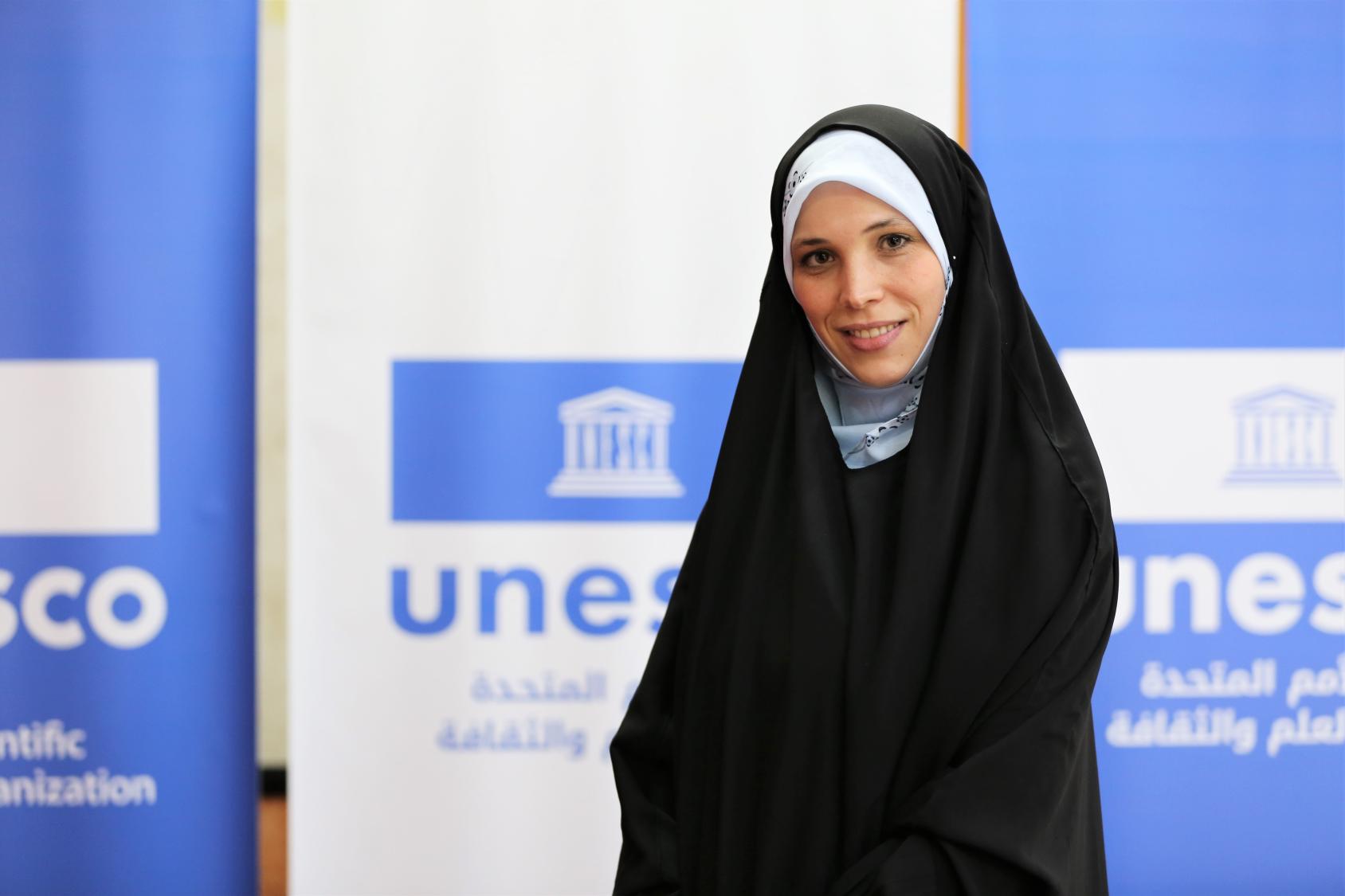 Una joven con un largo velo negro que le cubre el cuerpo y la cabeza posa sonriendo para la cámara. En el fondo hay grandes carteles azules y blancos con las palabras "UNESCO" debajo del logotipo de la organización.
