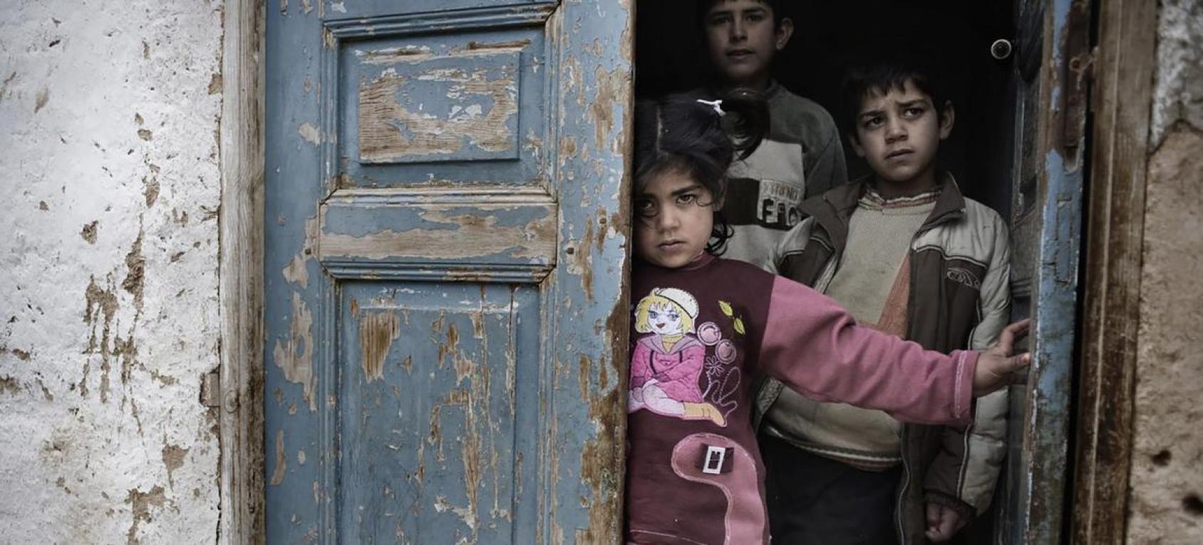 Une petite fille et deux petits garçons se tiennent debout, le regard triste, sur le pas d'une vieille porte bleue délabrée.