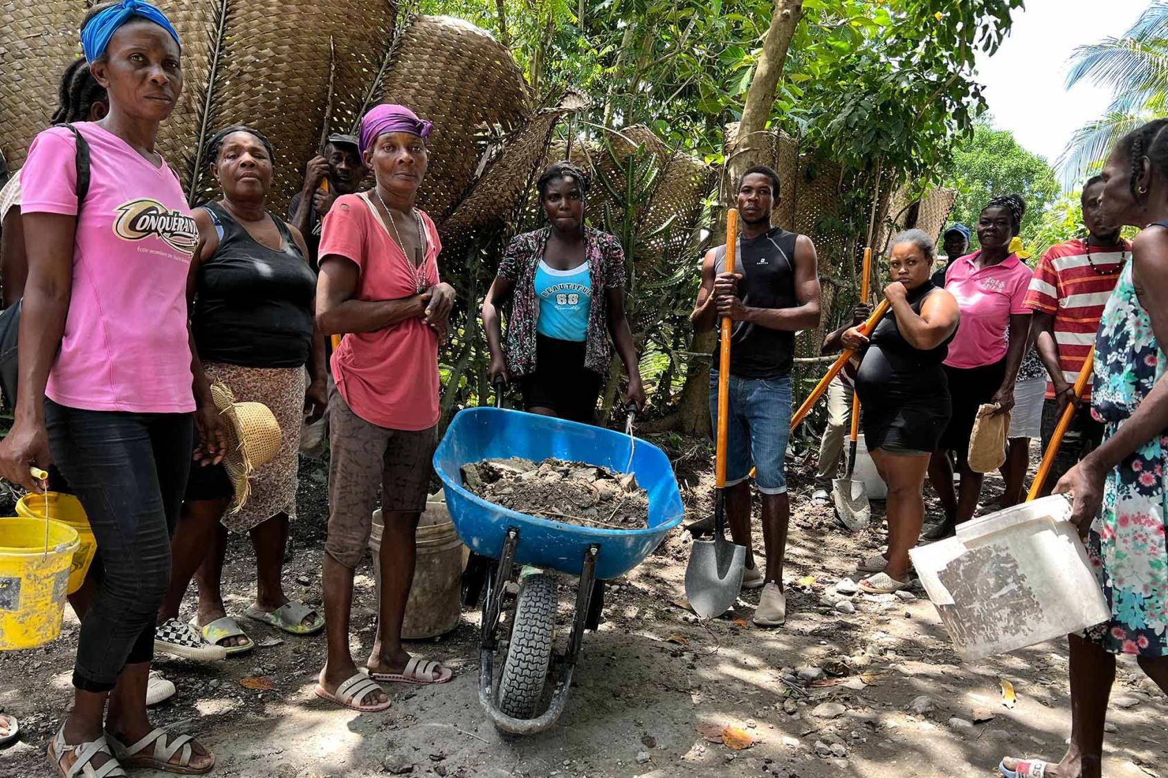 Un grupo de hombres y mujeres en Haití están de pie en un semicírculo en el exterior, sosteniendo varias herramientas y mirando con tristeza a la cámara. En el centro hay una carretilla azul.