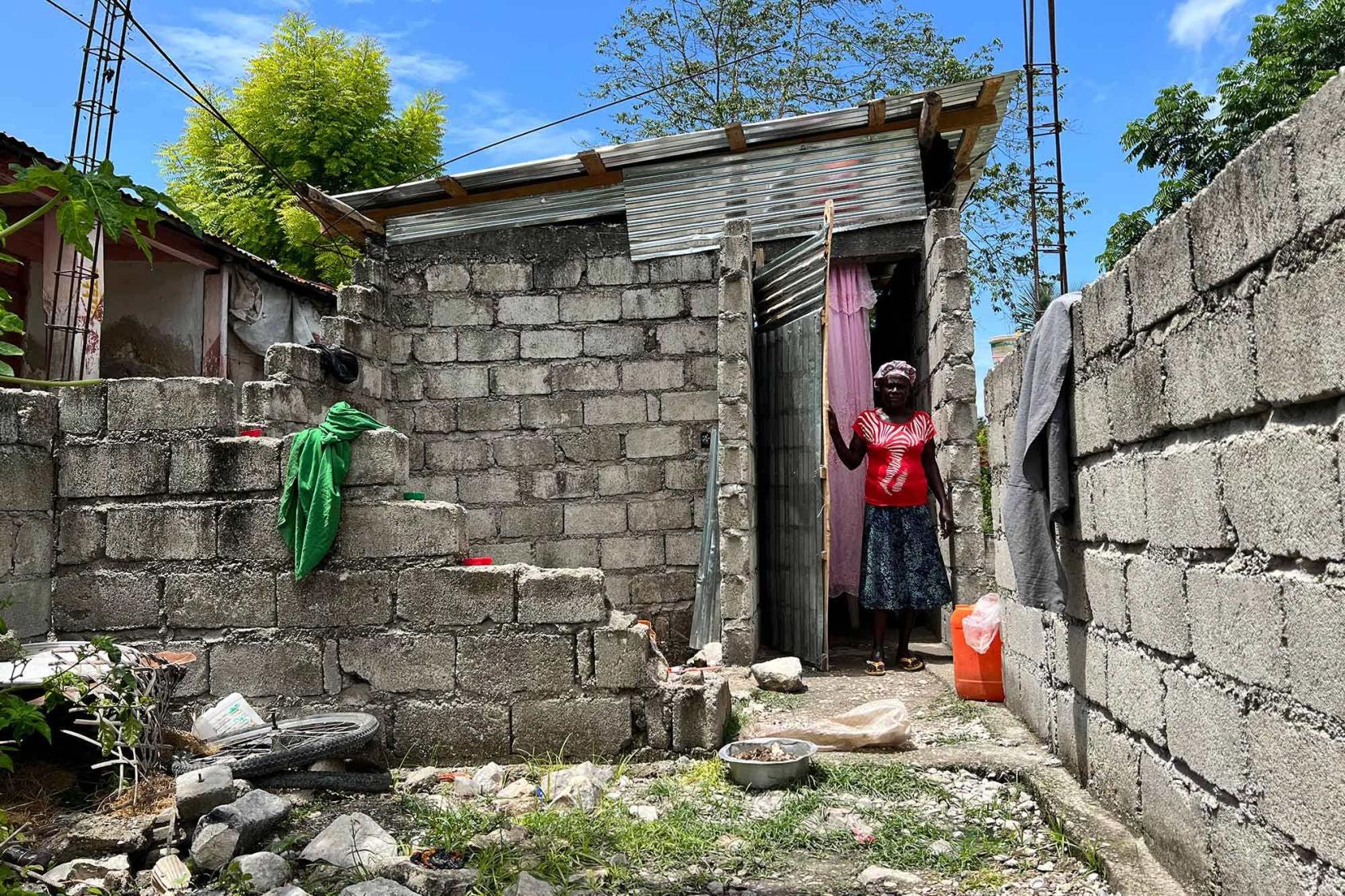 A Haitian woman stands in a doorway overlooking an outdoor courtyard enclosed by unpainted walls.