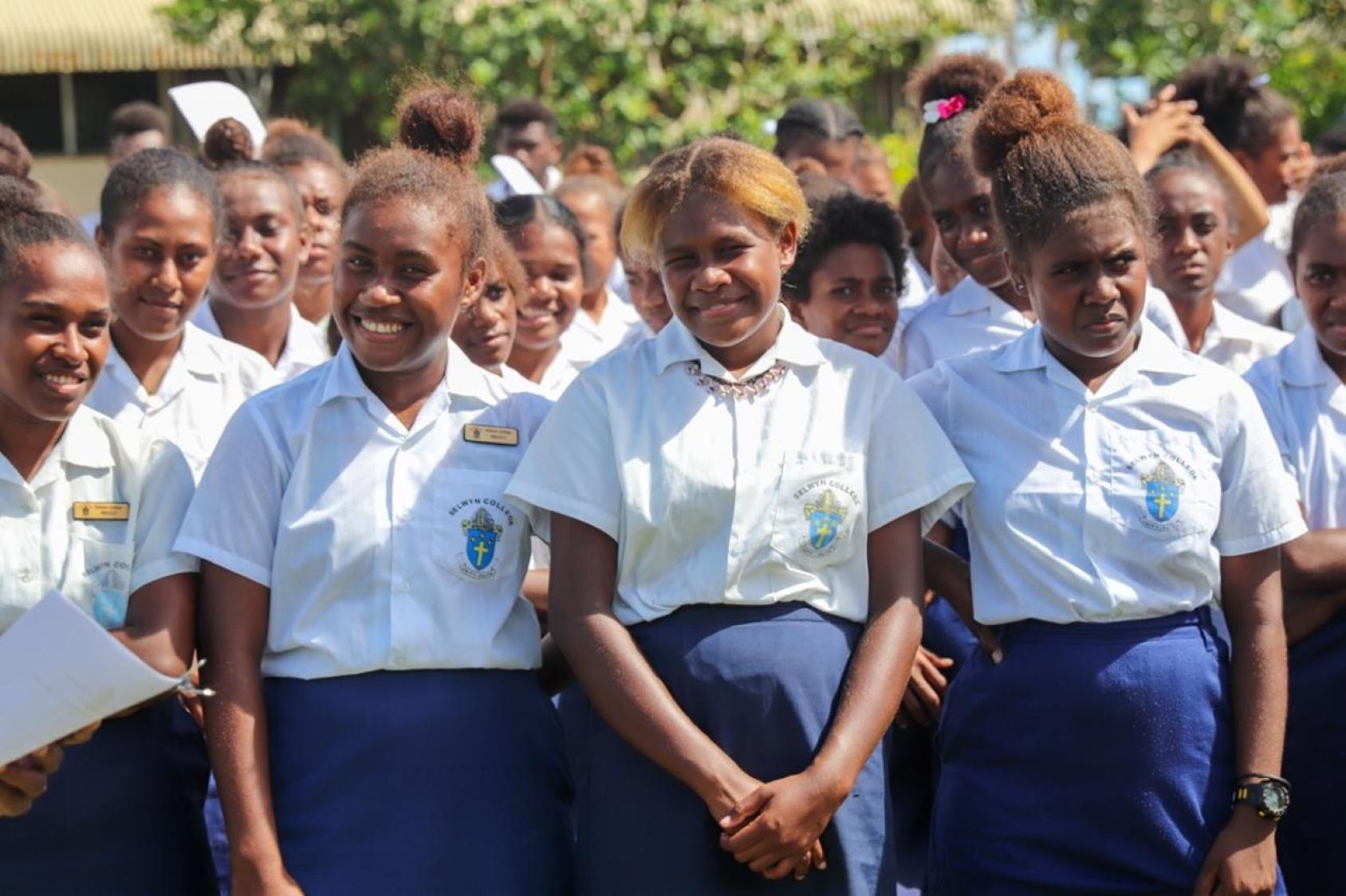 Aux Îles Salomon, des adolescentes en uniforme scolaire vêtues de jupes bleues et de chemises blanches posent devant la caméra en souriant. 