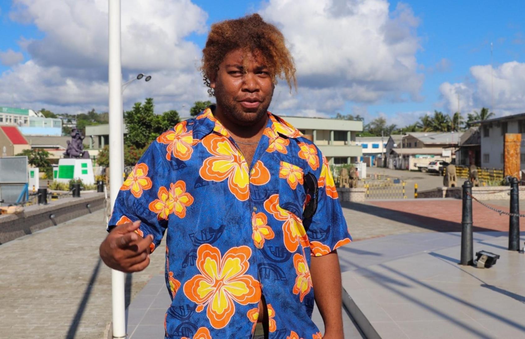 Aux Îles Salomon, un homme portant une chemise bleue imprimée de fleurs jaunes s'adresse à la caméra, en extérieur, par une journée ensoleillée. 
