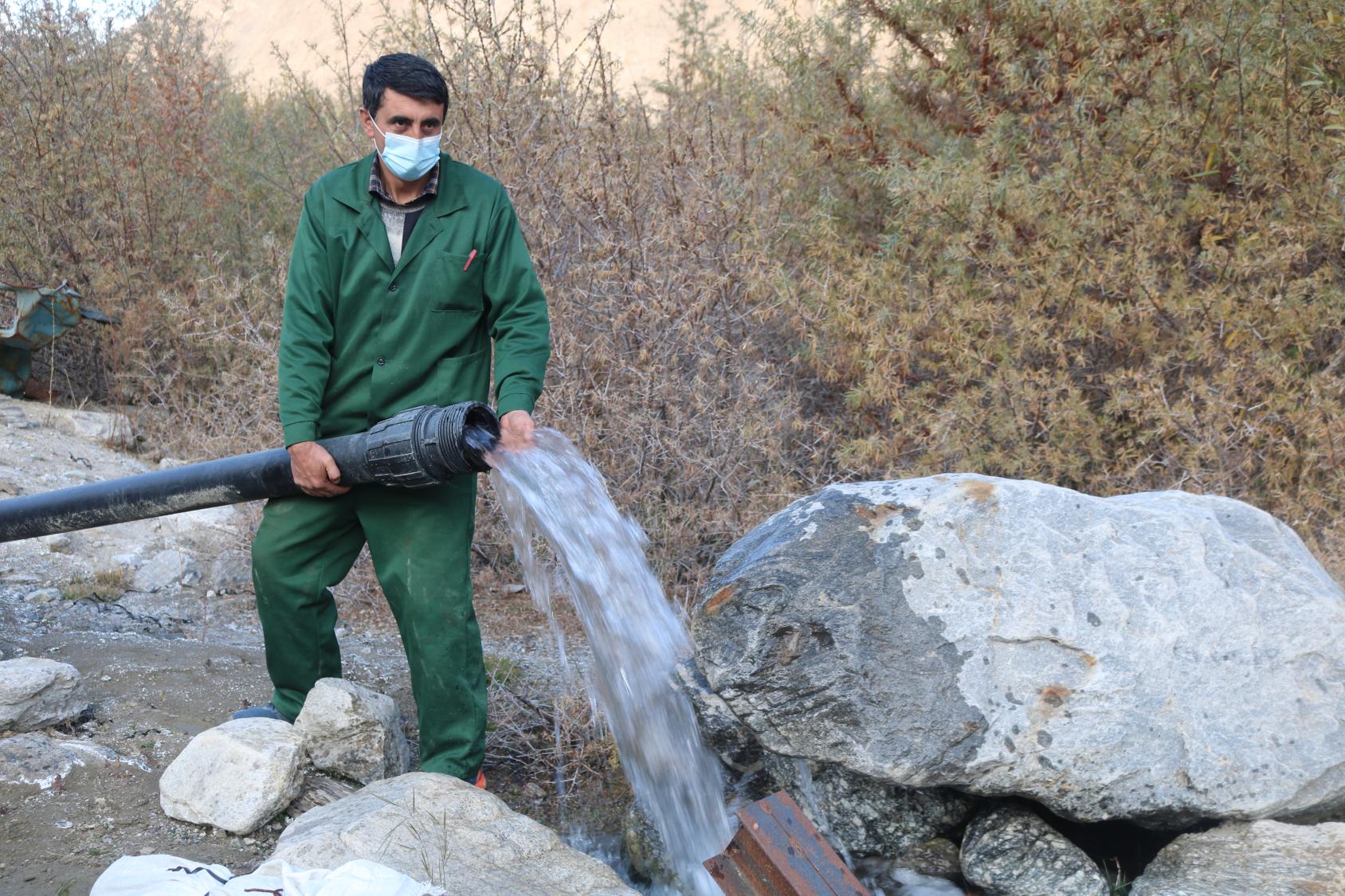 Au Tadjikistan, un homme en tenue de travail verte portant un masque de protection respiratoire tient un gros tuyau d'où jaillit de l'eau se déversant entre des rochers.