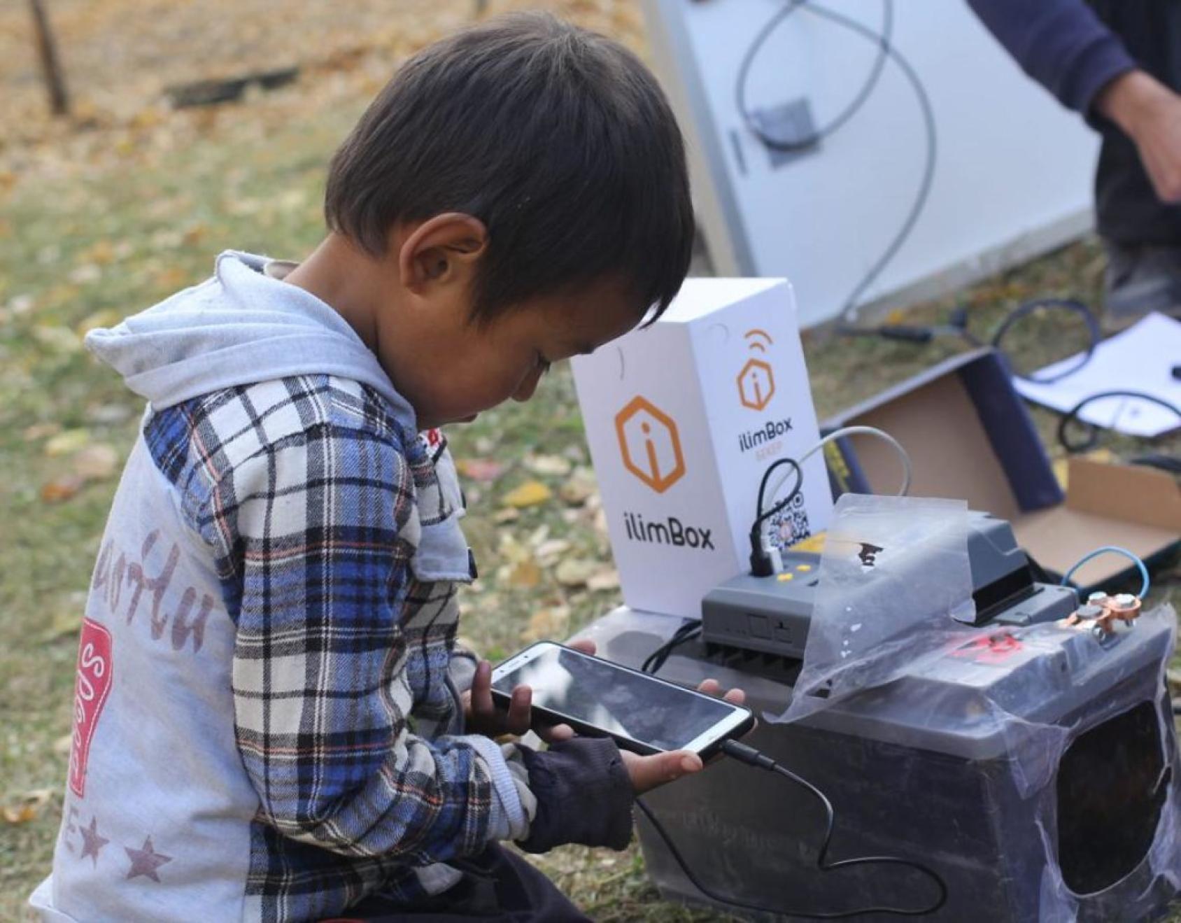 Au Kirghizistan, un petit garçon lit l'écran d'un téléphone portable connecté via un câble au dispositif "Internet in a Box" (en français : "Internet dans une Boxe"), qui permet de stocker des contenus dans une bibliothèque numérique.