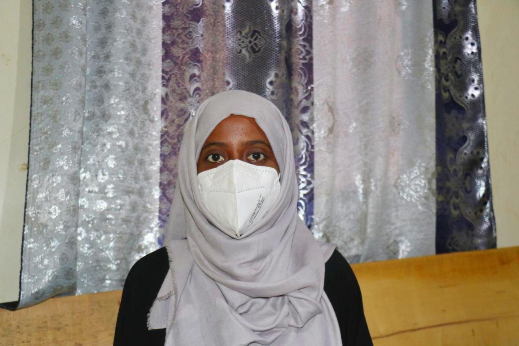 Photo-portait d’une jeune femme yéménite portant un voile gris autour de la tête et des épaules et un masque de protection respiratoire sur la bouche et le nez.