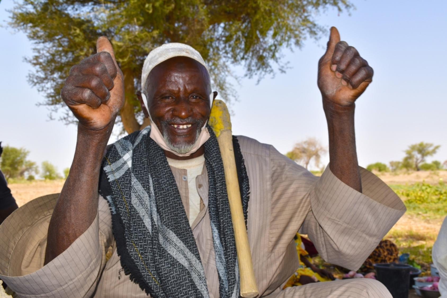 Un homme camerounais portant un vêtement traditionnel beige, un couvre-chef blanc et une longue écharpe grise autour du cou est assis en tailleur au milieu d'une étendue verdoyante et lève les deux pouces vers le ciel en regardant la caméra avec un large sourire.