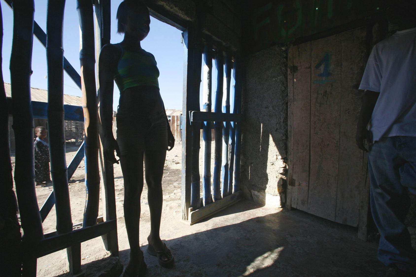 A young woman is photographed in backlight standing on the threshold of an entrance made of wooden bars.