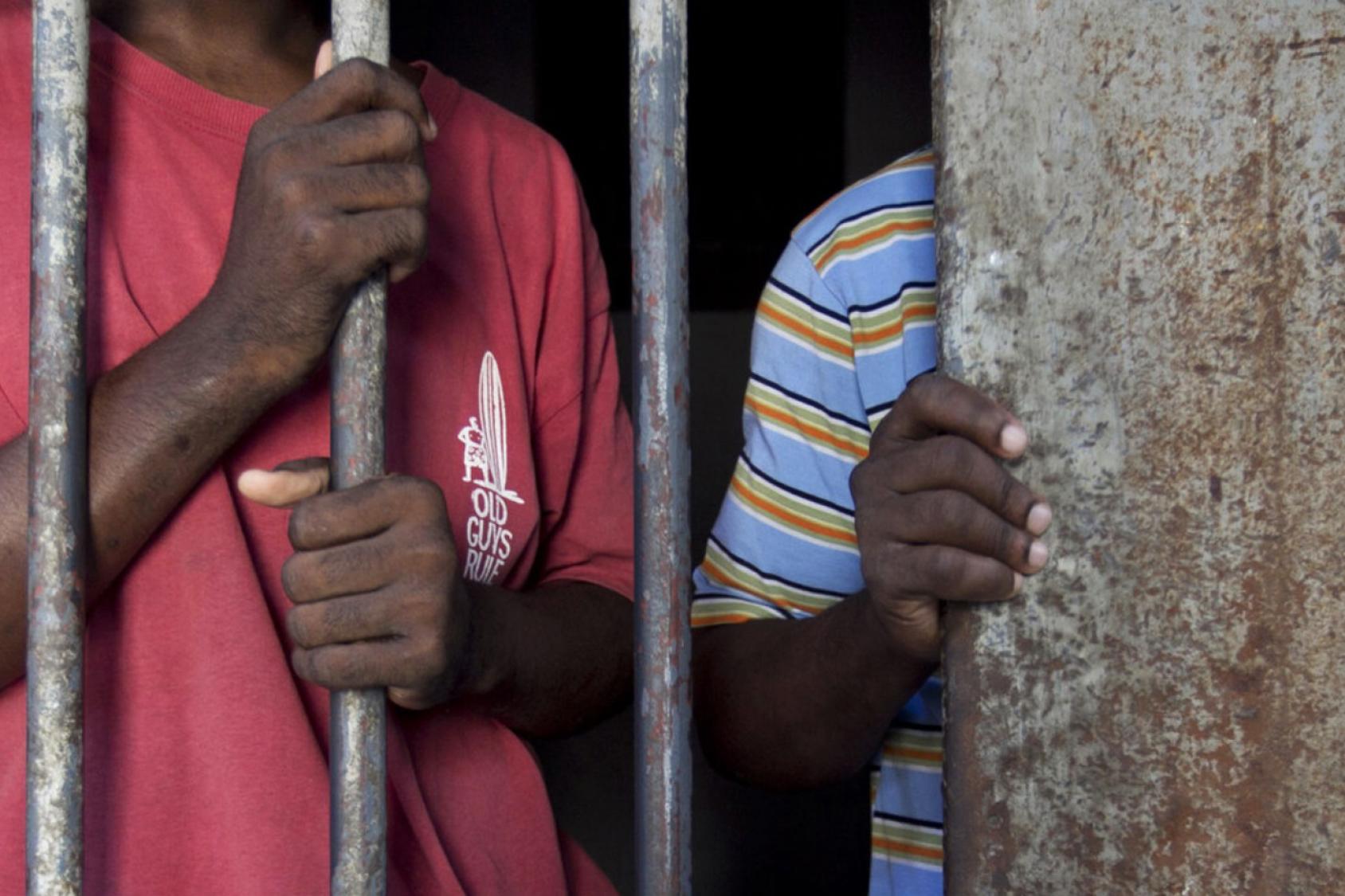 Close-up of two boys with their faces off-camera standing behind the bars of a prison cell.
