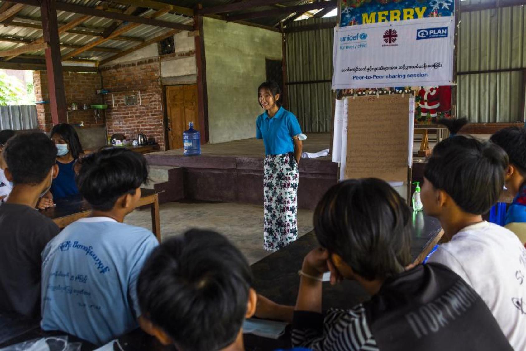 A young woman standing in front of some teenagers, speaking to them in a peer-to-peer knowledge sharing session.