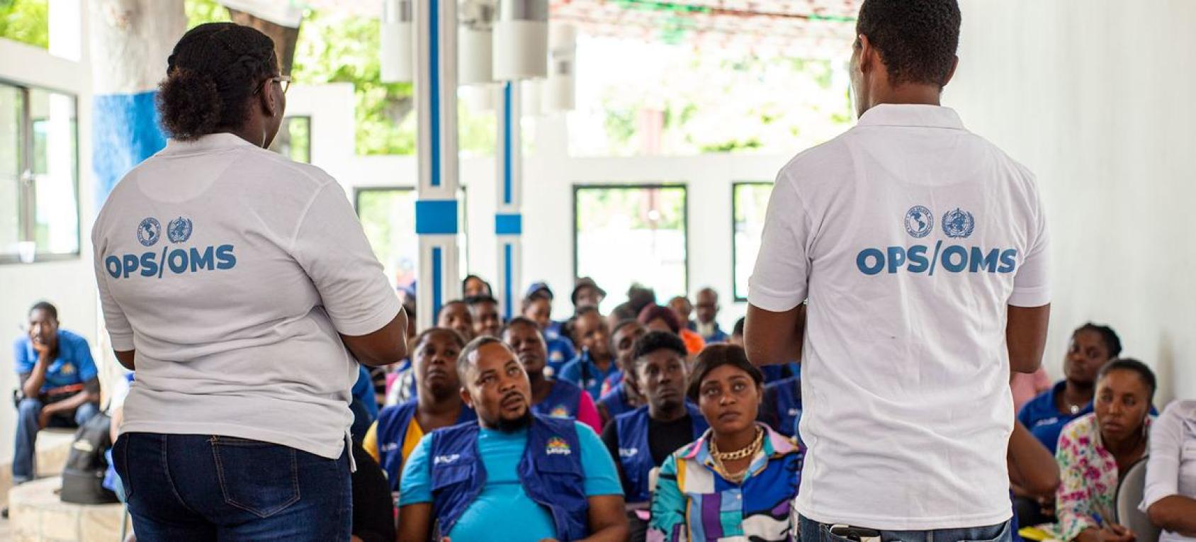In Haiti, a man and a woman wearing a white T-shirt with "PAHO/WHO" written on it provide cholera revention training to health care workers sitting in a room. 