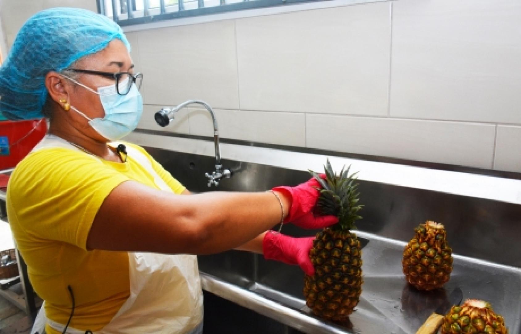 Une femme portant un masque de protection respiratoire, des gants, une charlotte et une blouse jaune, nettoie un ananas dans un évier industriel.