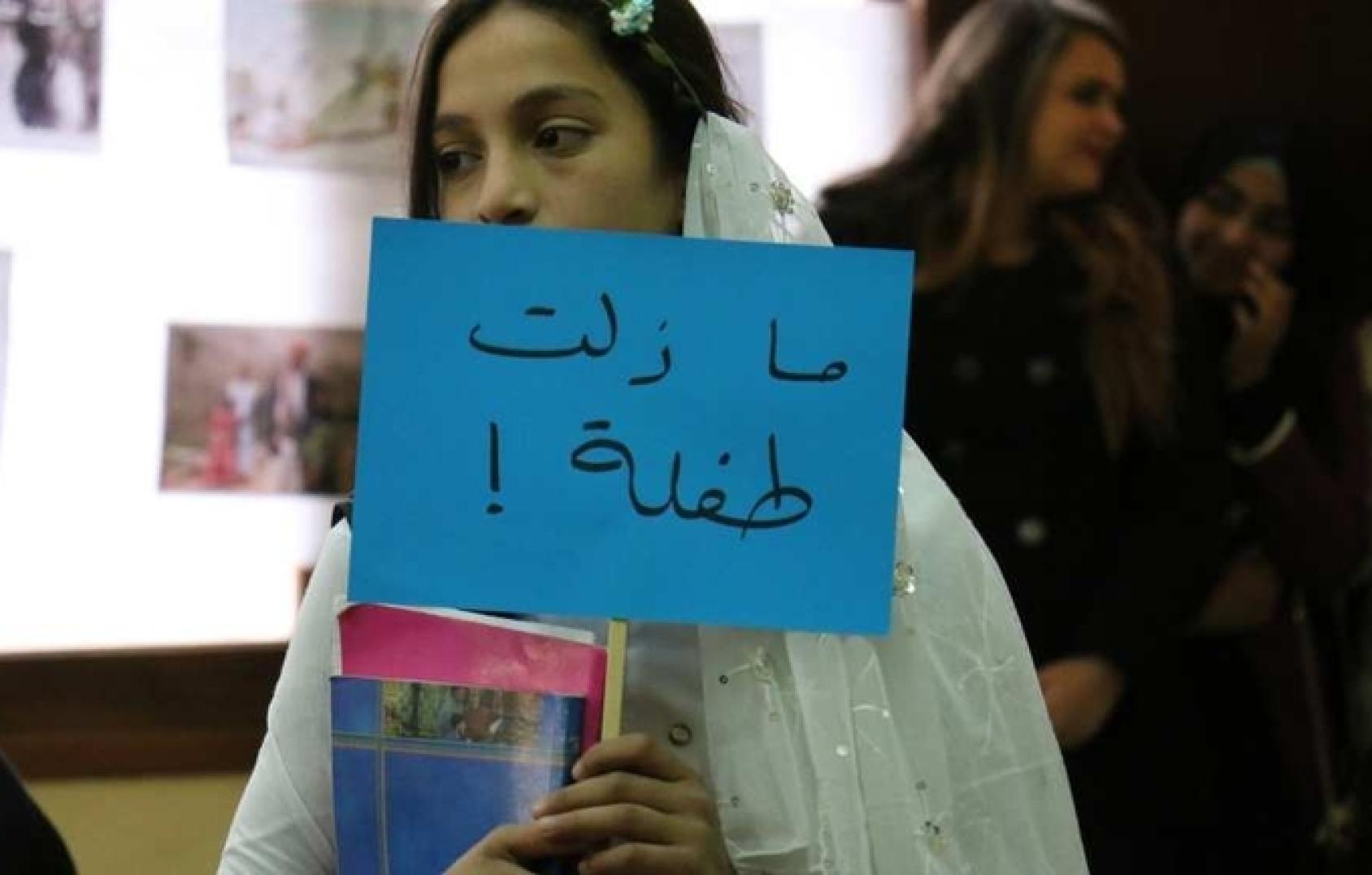 A young girl wearing a wedding dress and holding a sign that says: I am still a child.