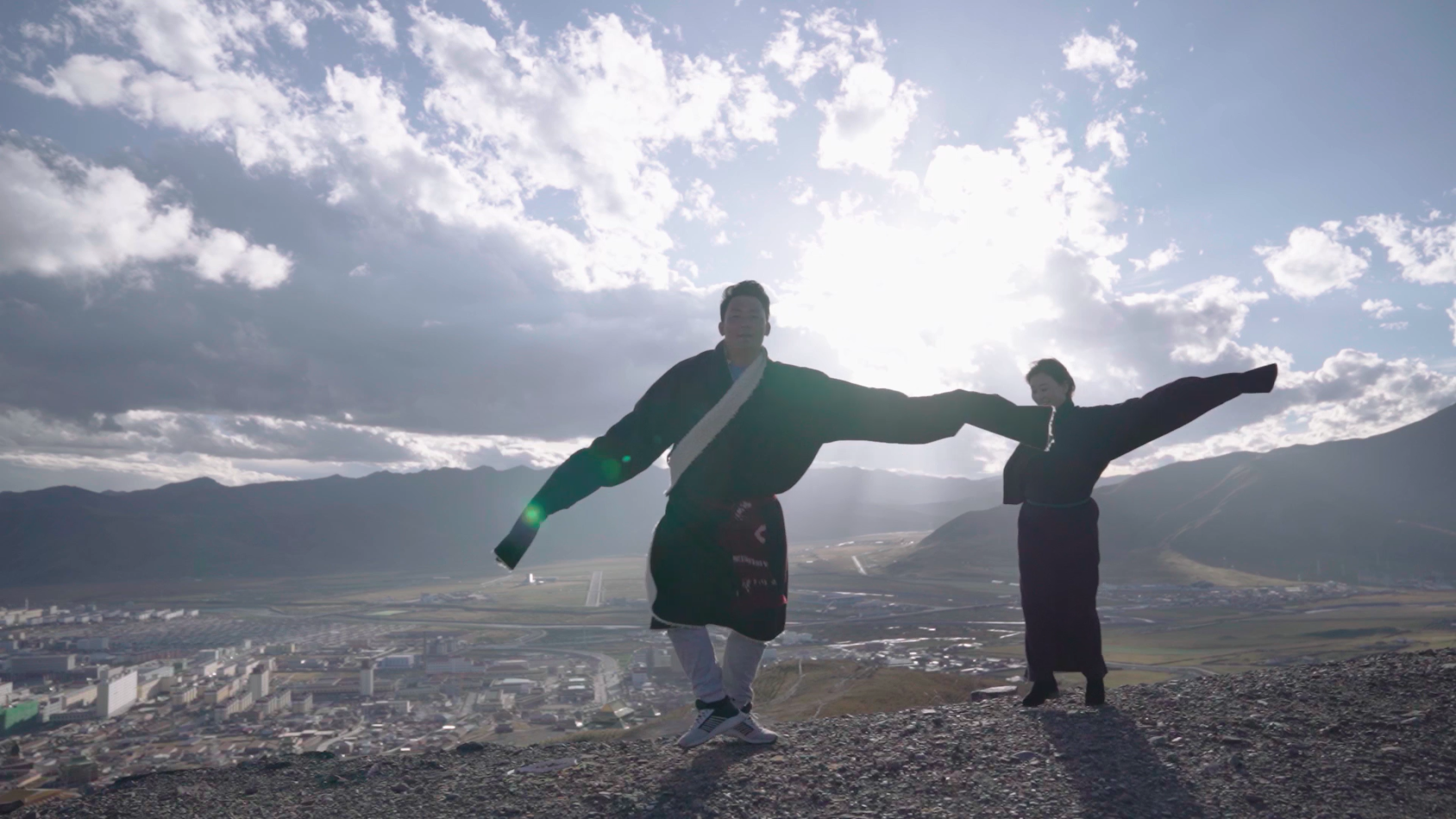 Dos personas tibetanas bailando en una meseta con el fondo del cielo iluminado por el sol. 