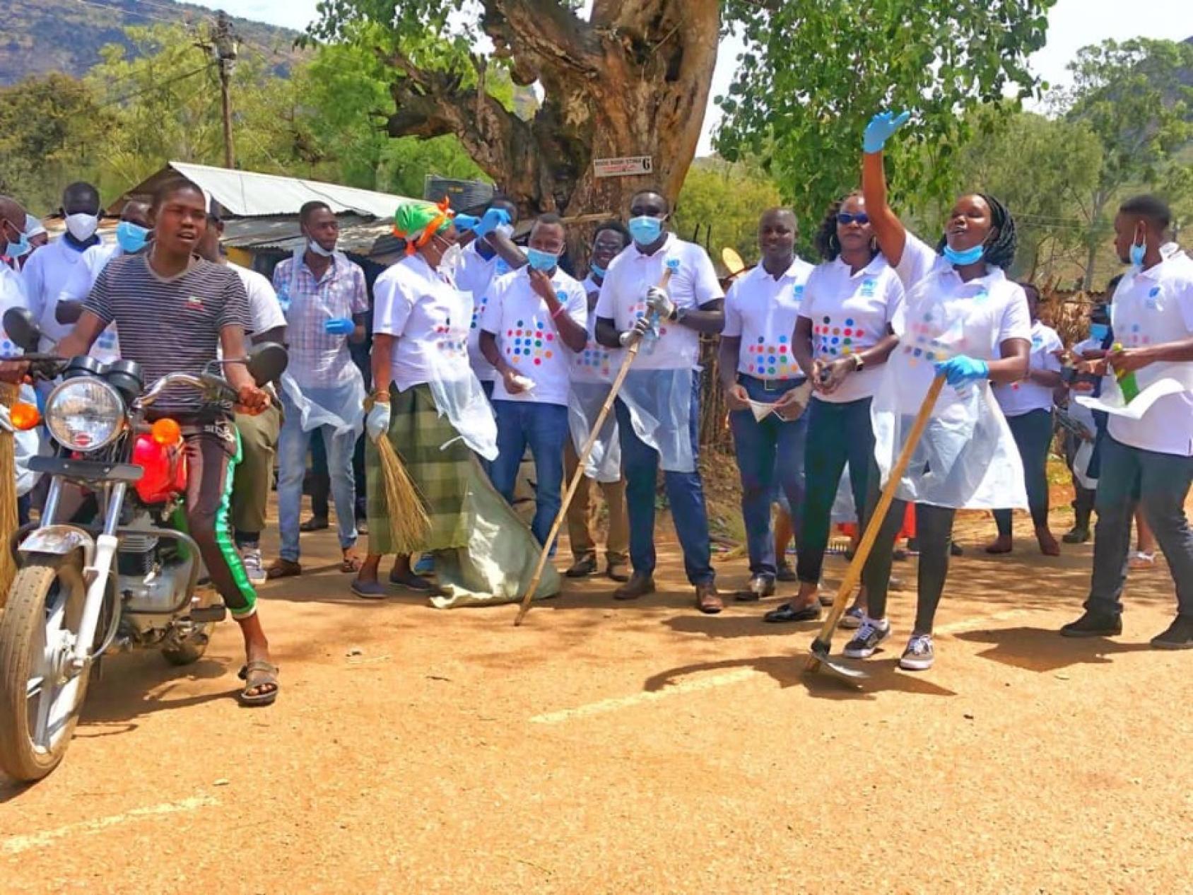 On a sunny day, a group wearing UN Uganda (SDG) t-shirts, appear to be cleaning and smiling.