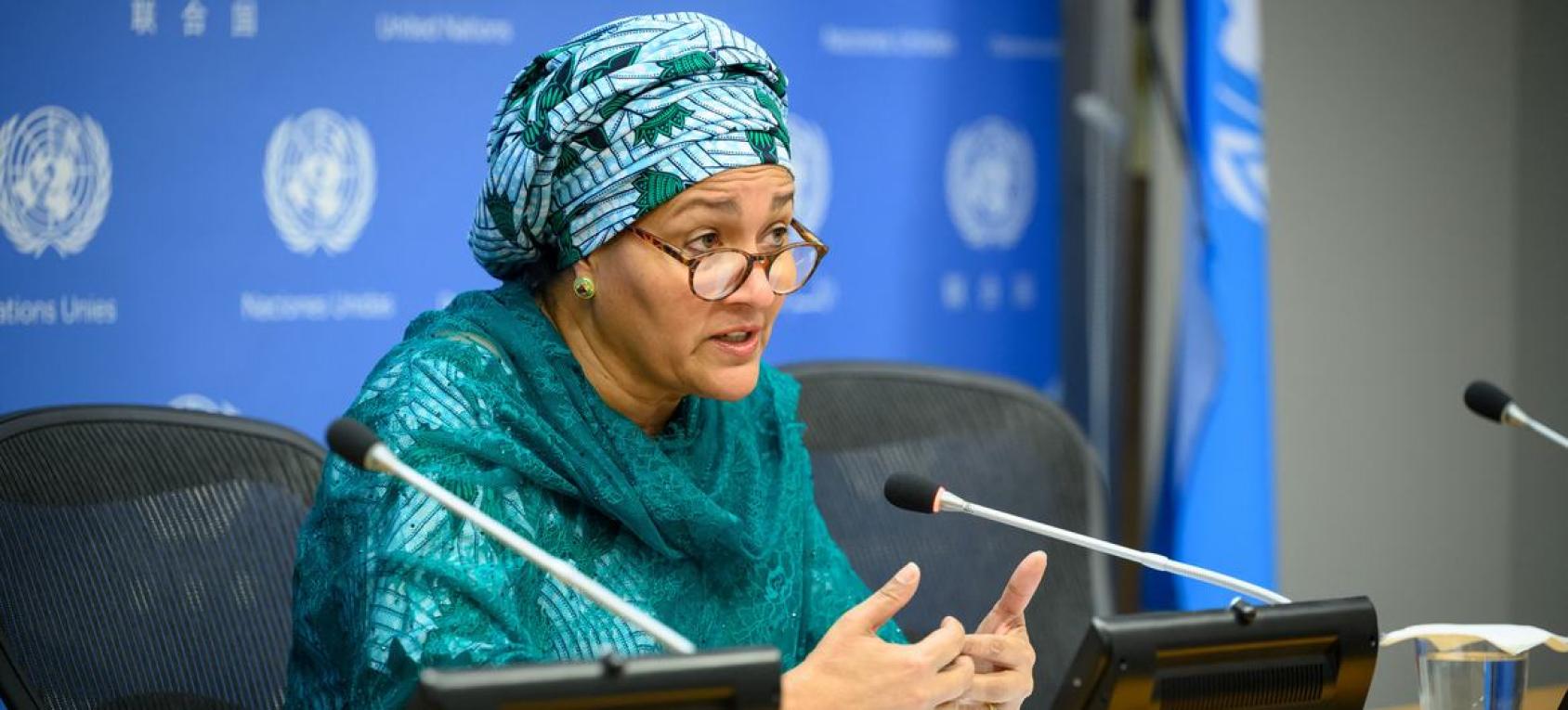 Woman in green dress and headscarf speaking into a microphone against a blue background with UN logo