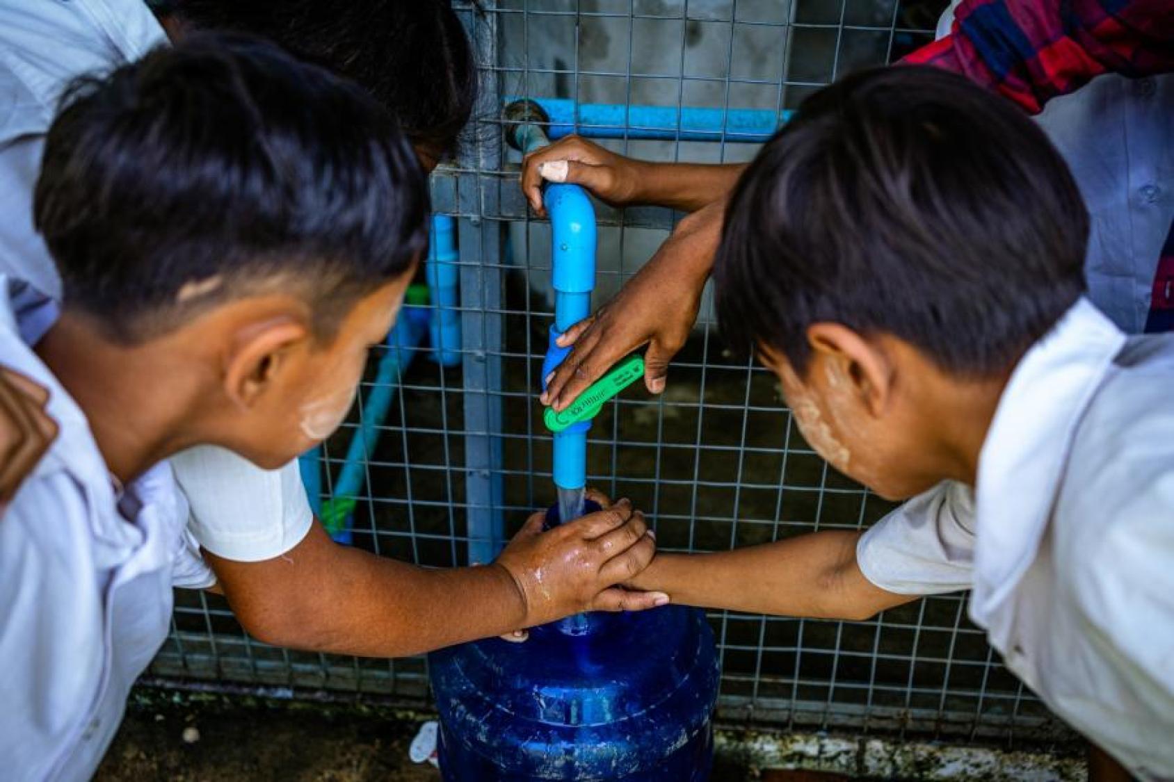Boys, wearing white polo shirts, fill a container with water from a drinking fountain.