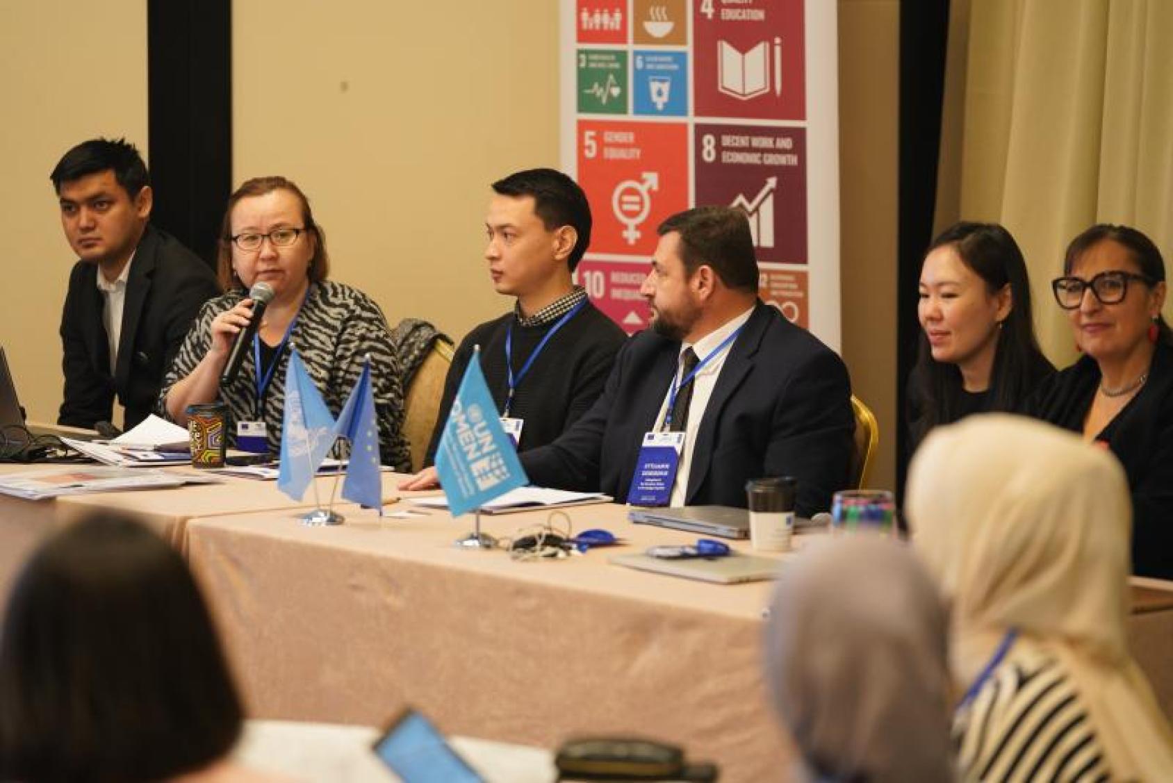 A panel of suited speakers discuss topics, with an SDGs banner behind them and UN Women flags on the table.