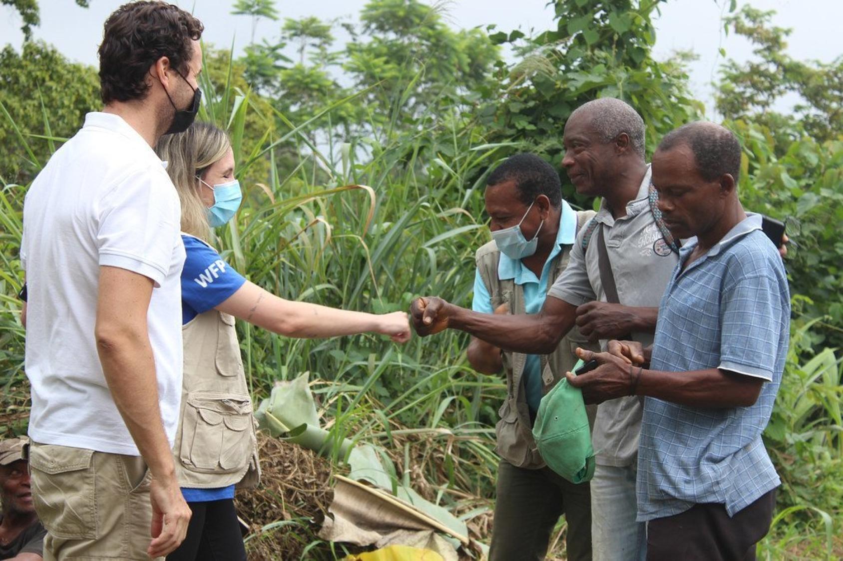 A few men, who appear to look like farmers, talk to a woman in a WFP vest and a man, in a lush green location.