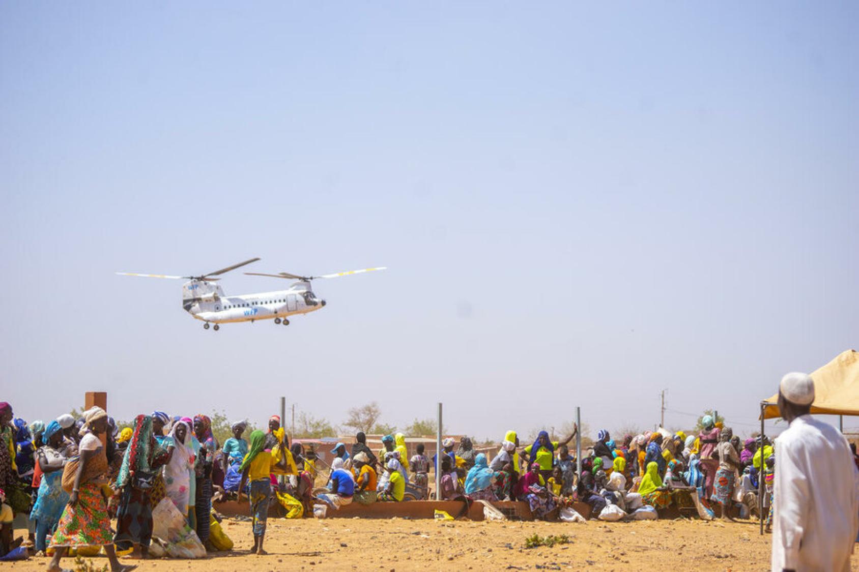 A low propeller style plane flies over a dry land with groups of people in colourful clothes gathered