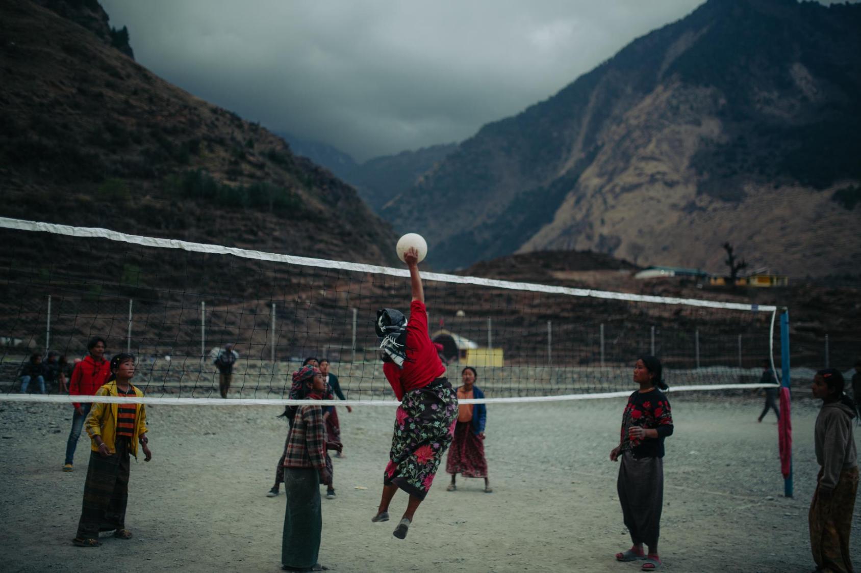 Against mountains in the background, a group of women in colourful red clothes play volleyball over a net in Nepal