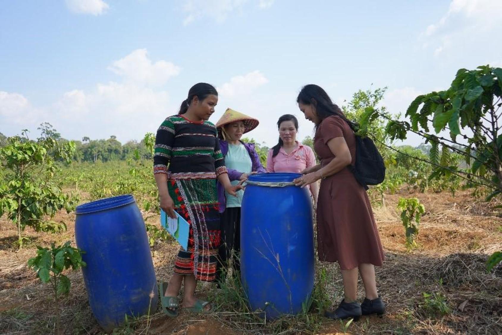 Four women in skirts and straw hats standing over two big blue vats against a background of trees and soil