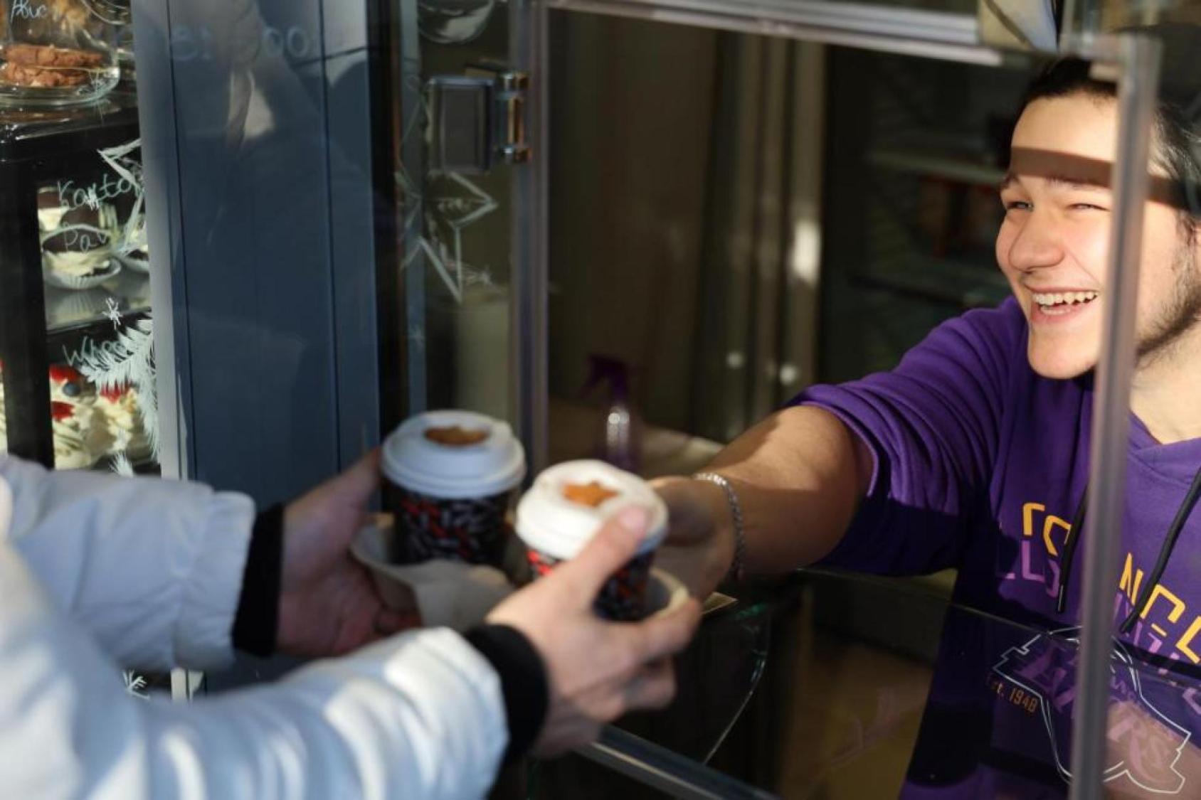 A man in a purple shirt smiles as he hands a cup of coffee to a customer in a kiosk