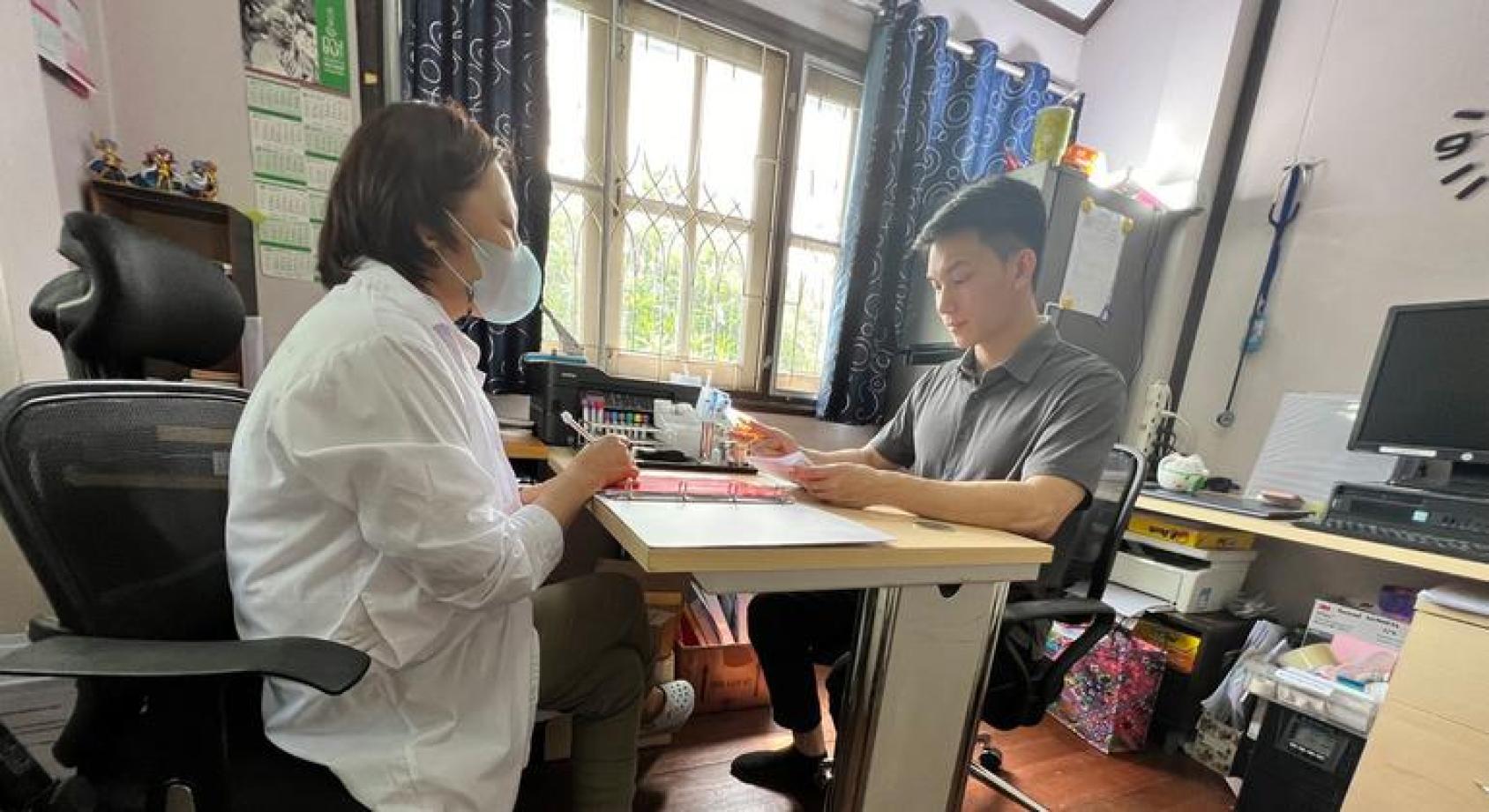 A woman in a white coat and a face mask like a doctor sits across the table from a man in a grey shirt in a clinic