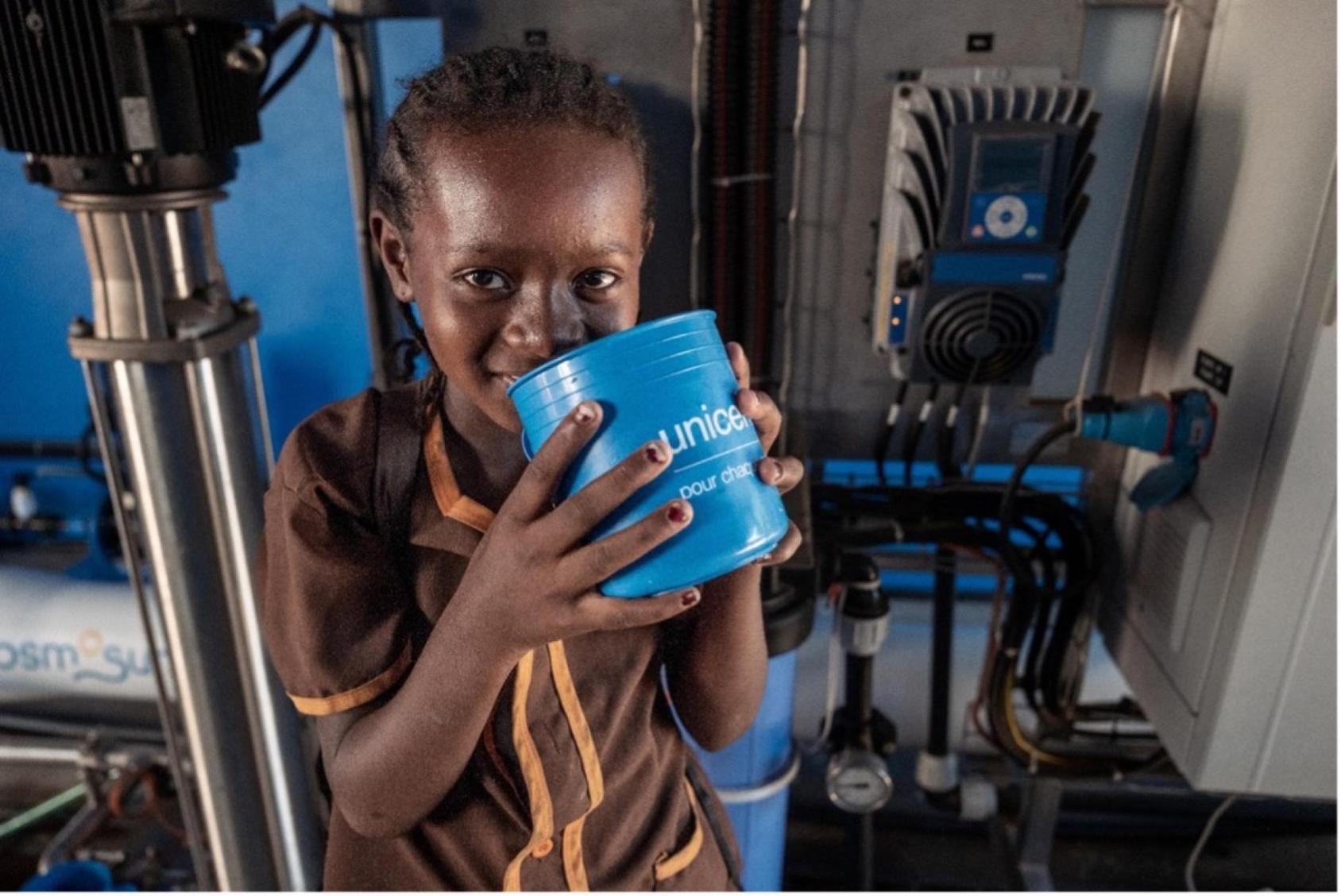 Child drinks water from a blue tumbler with a UNICEF logo in a room with a purification machine