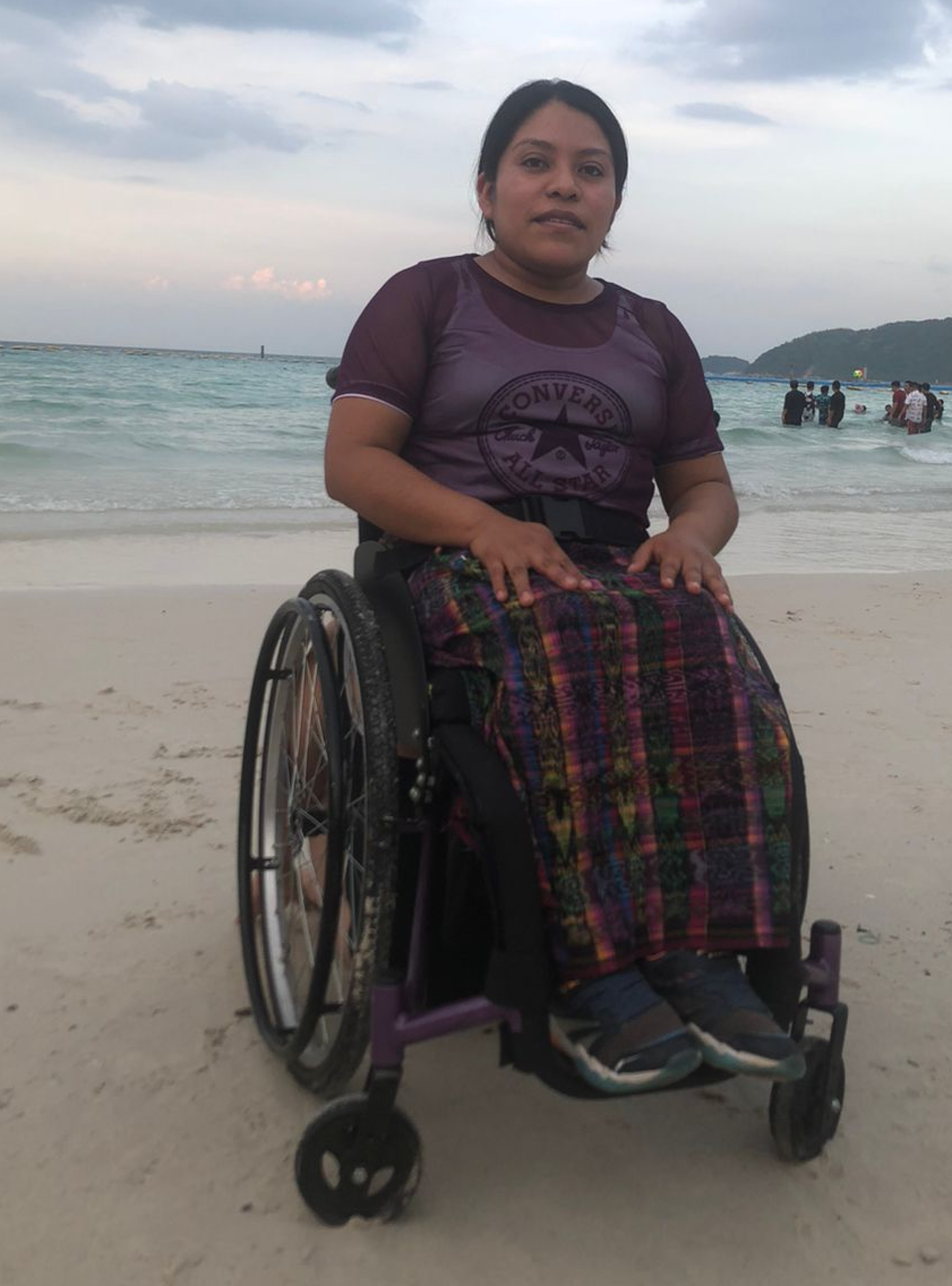 A woman in a brown shirt and pants sits on a wheelchair on a sandy beach in Guatemala