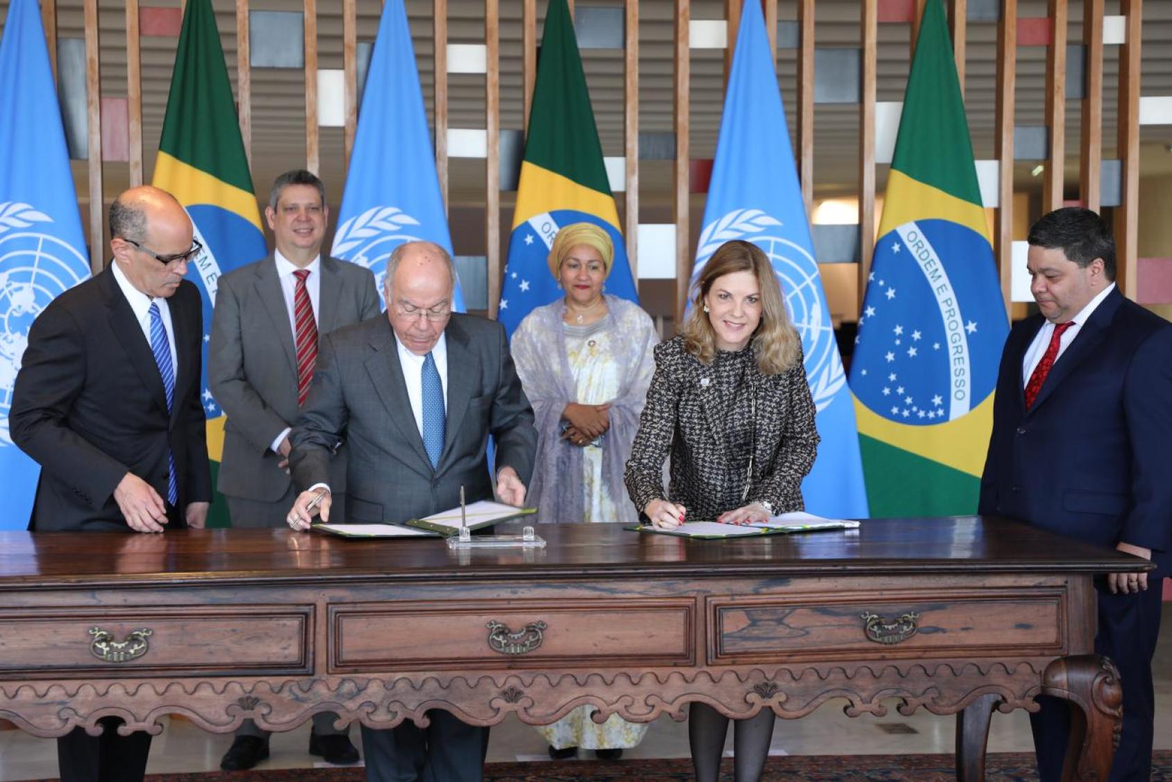 Four men and two women stand around a table where a man and a woman sign a document in front of the flags of UN and Brazil