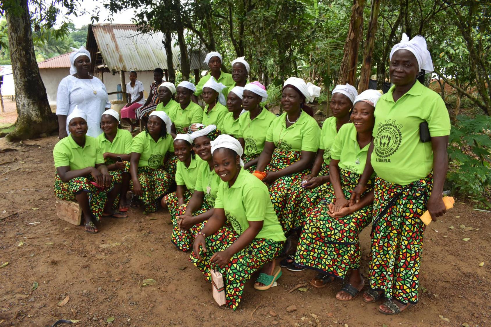 A group of women in green shirts and skirts and white headscarves smile at the camera