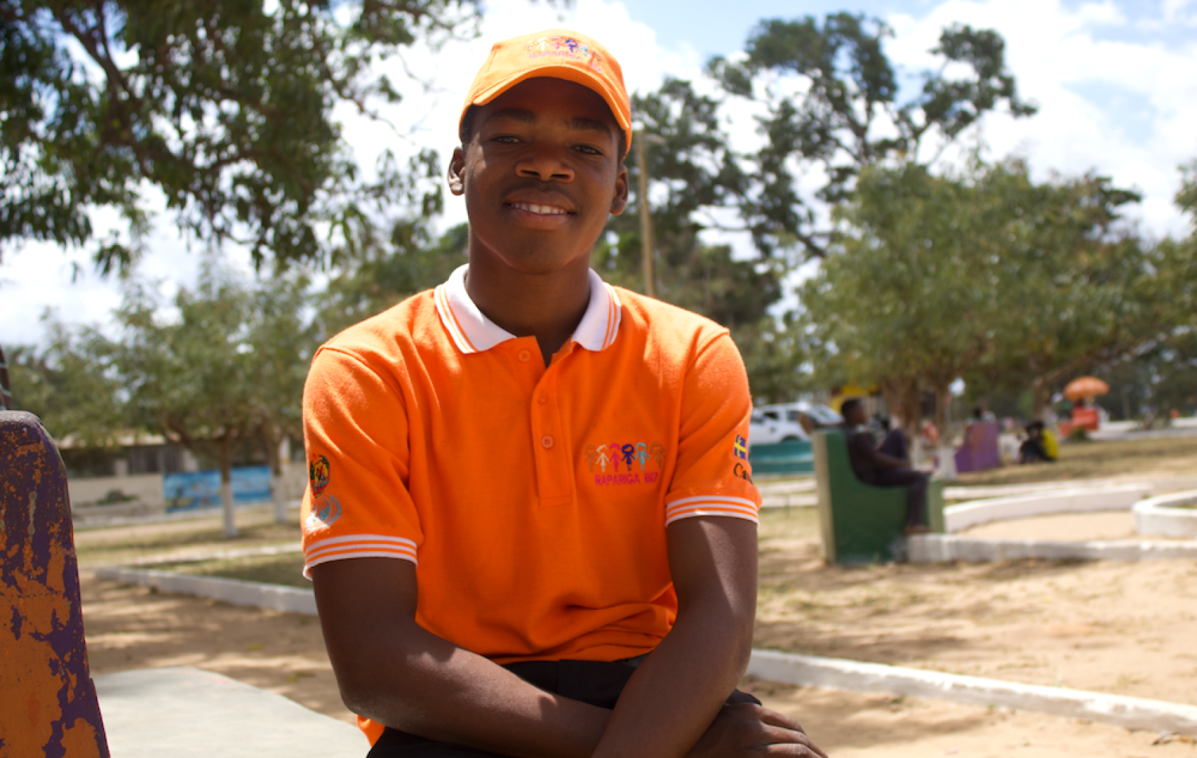 A young Mozambican man in an orange shirt and a cap sits in an open park