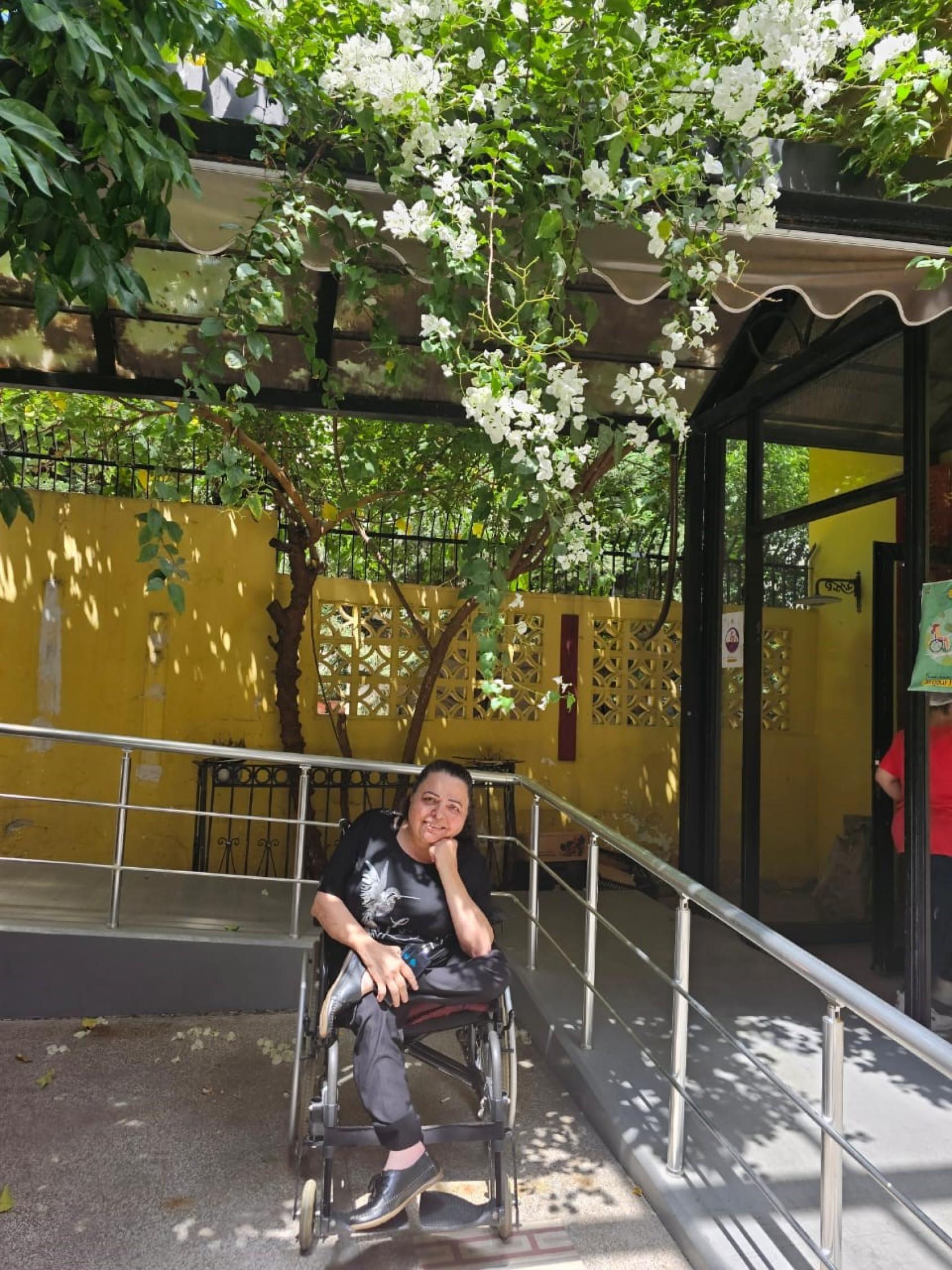 A woman in a wheelchair sits in a sunny courtyard painted yellow, with trees.
