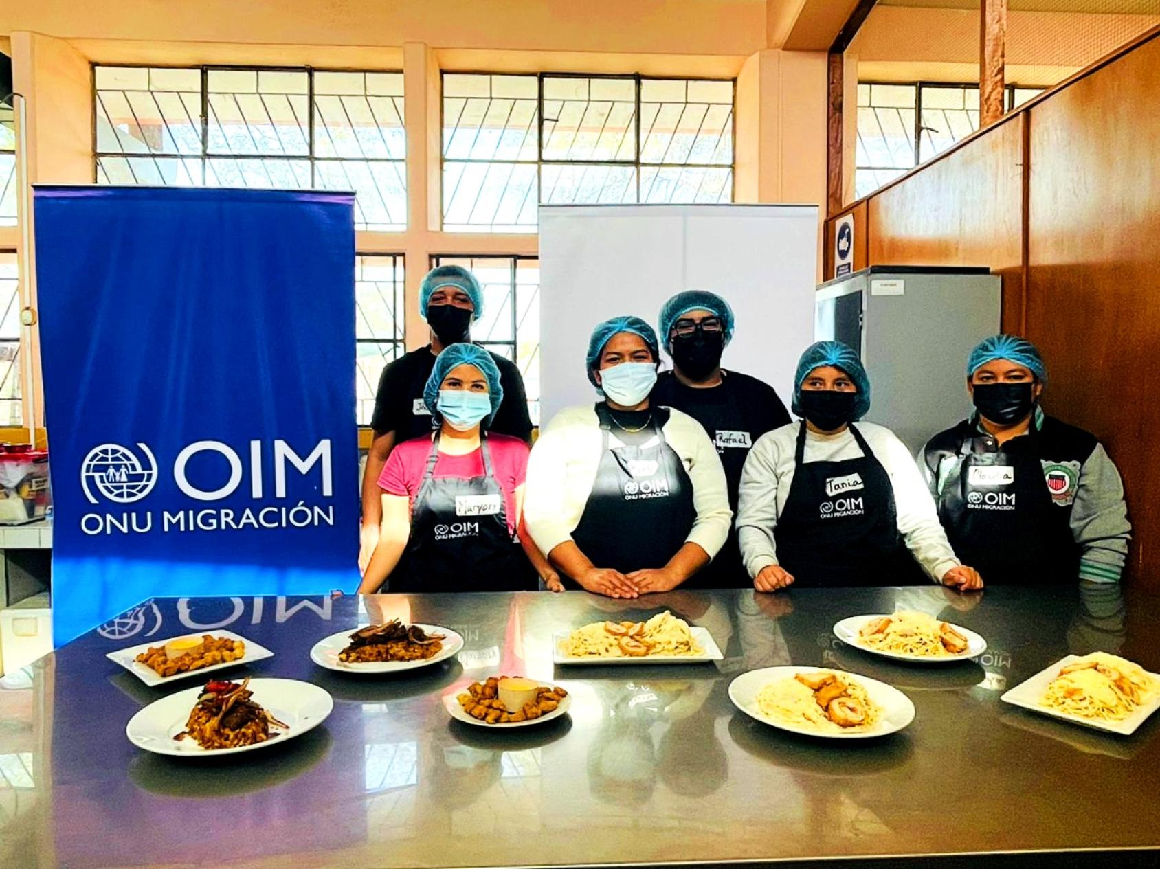 A group of six chefs stand at a table with their dishes displayed next to a blue IOM banner