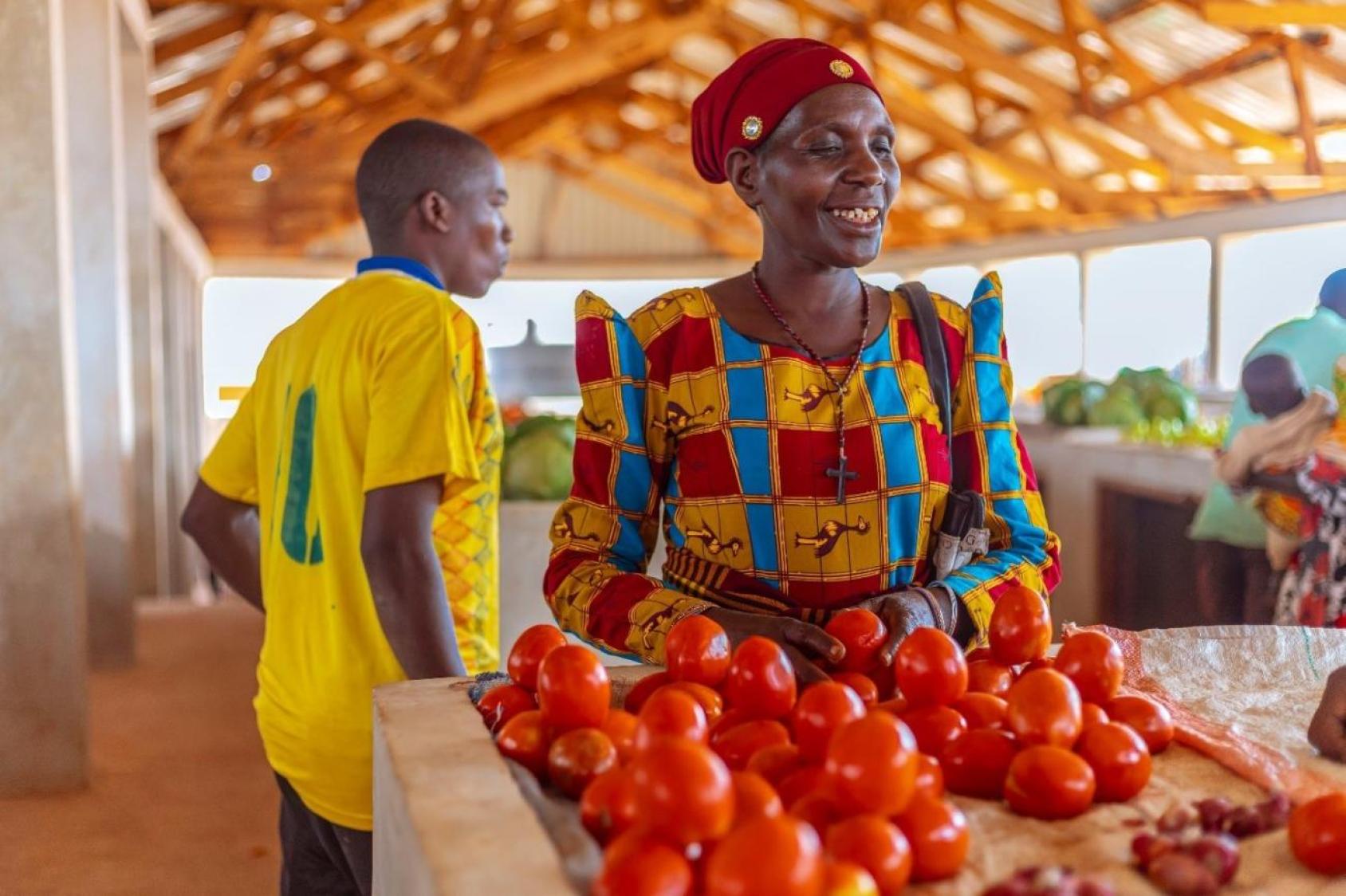 A woman in a red dress and headscarf and a man in a yellow sports jersey stand behind a makeshift stand with tomatoes